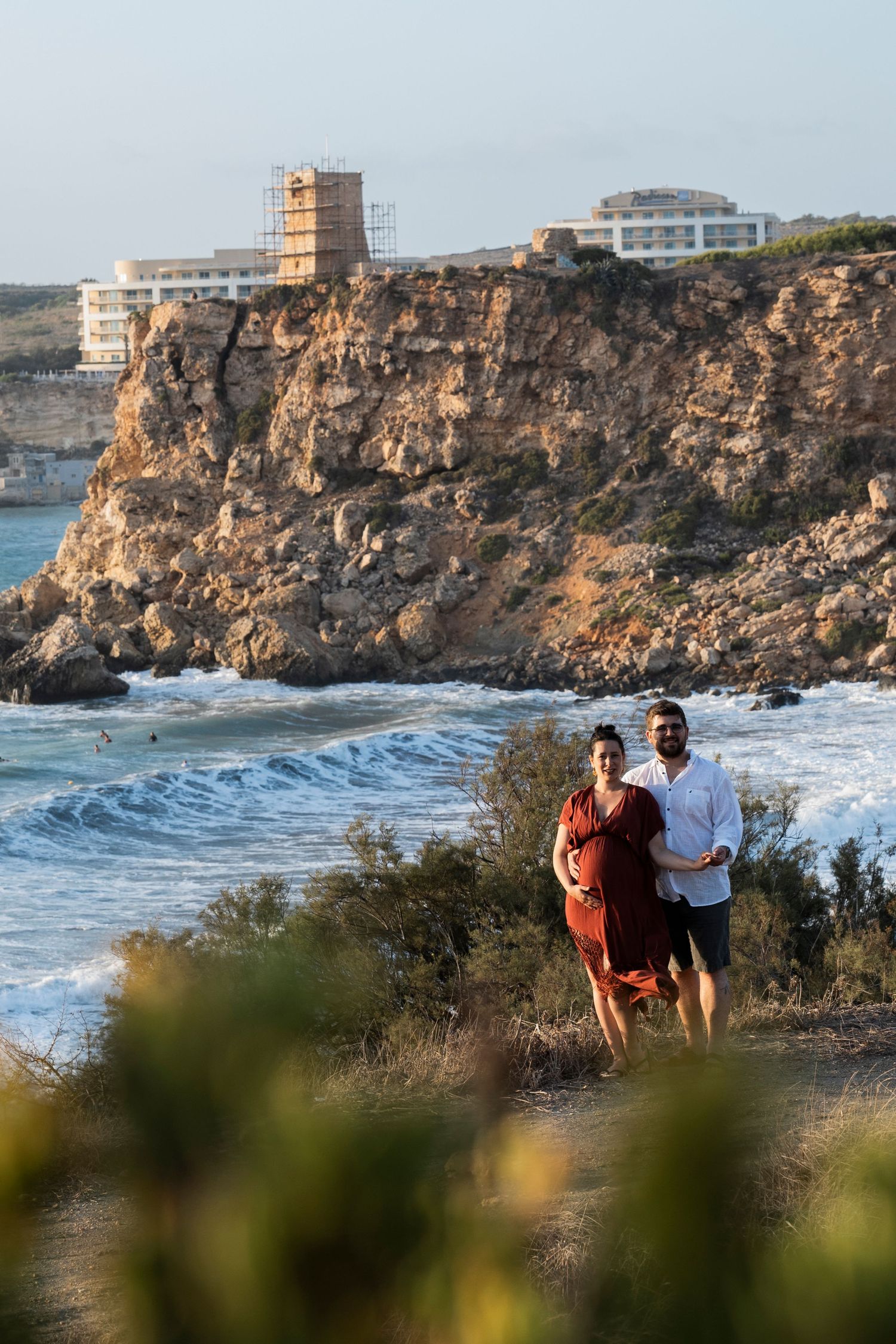 Couple embracing on rocky coastal cliffside with ocean waves and historic tower ruins in golden sunset light.