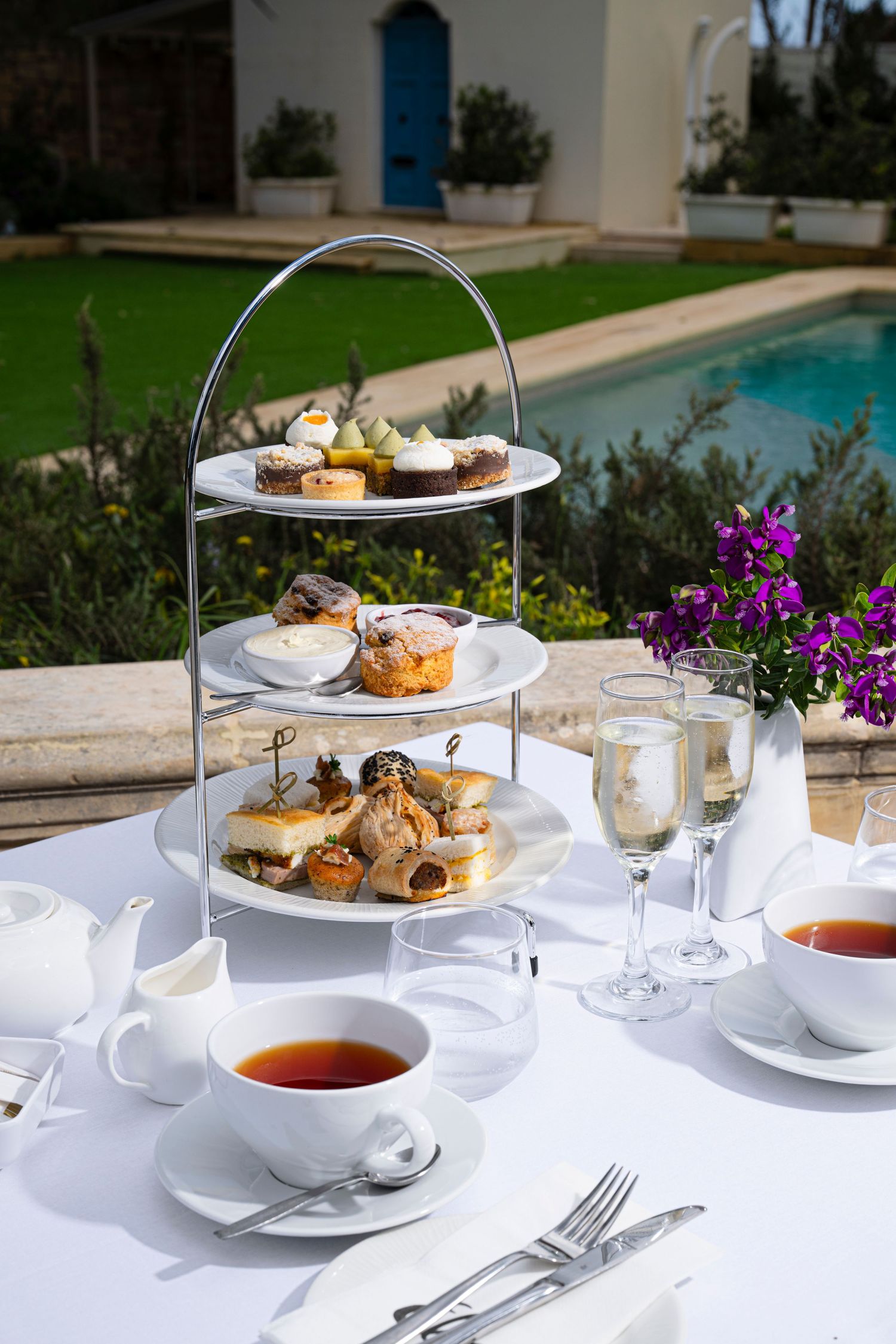 Elegant afternoon tea service with tiered stands of pastries and teacups set on white linens beside a swimming pool.