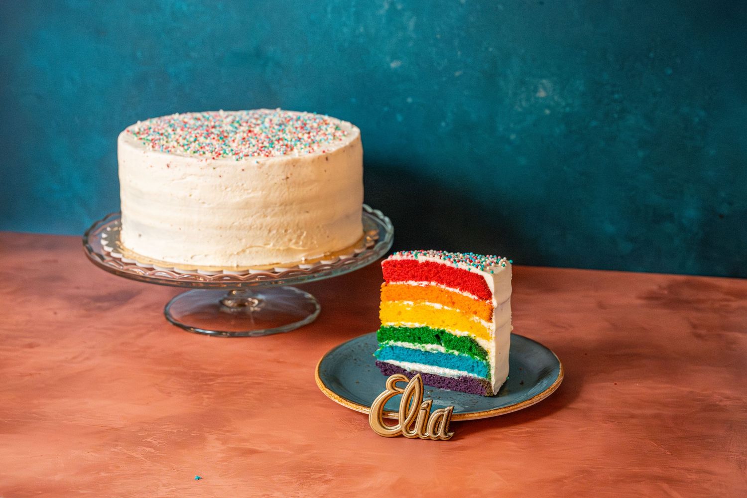 Rainbow layer cake slices displayed on plates against a teal background with gold decorative elements.