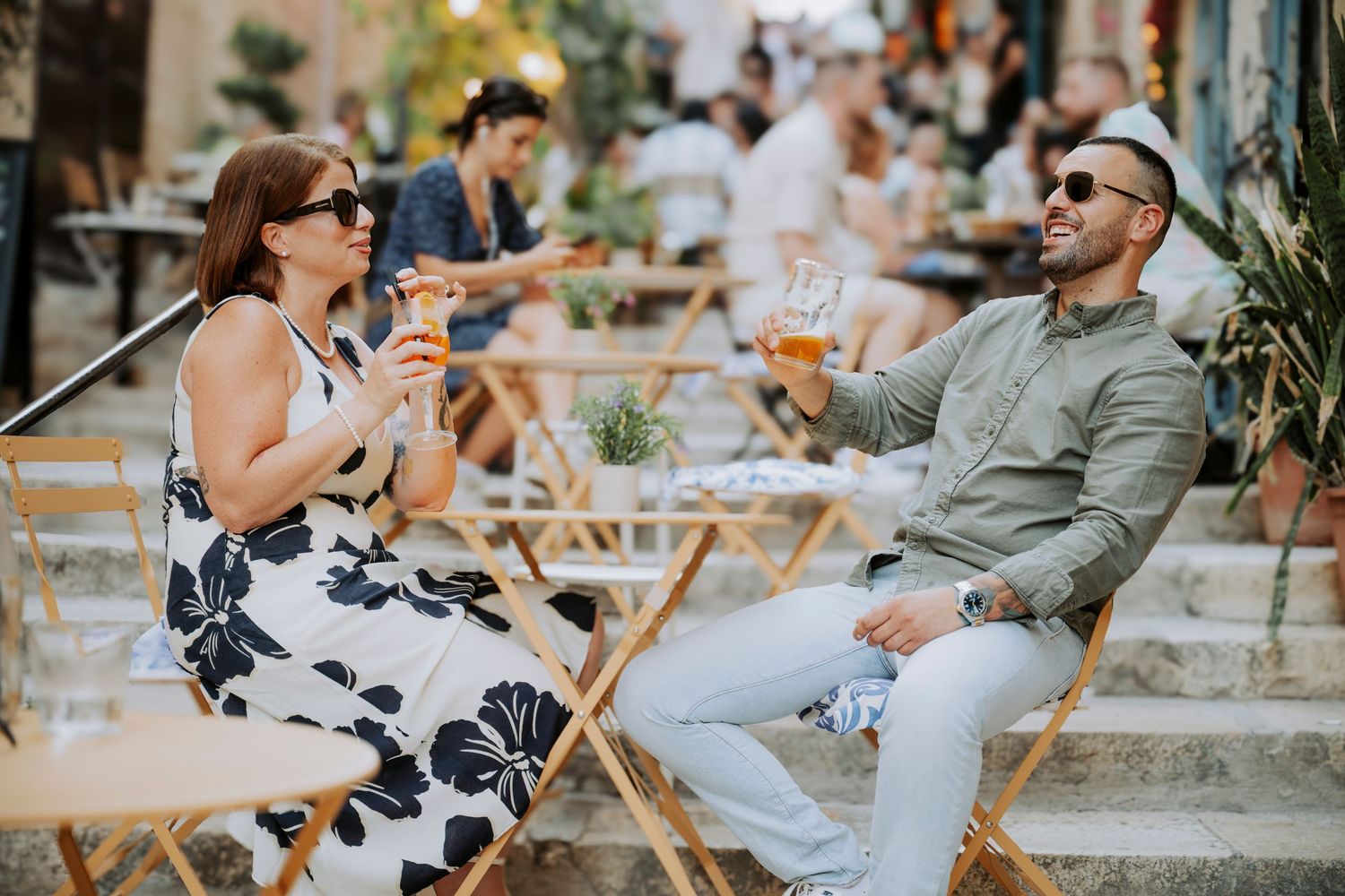 Couple enjoying drinks at an outdoor cafe patio, woman in floral dress and man in white cap smiling on sunny day.