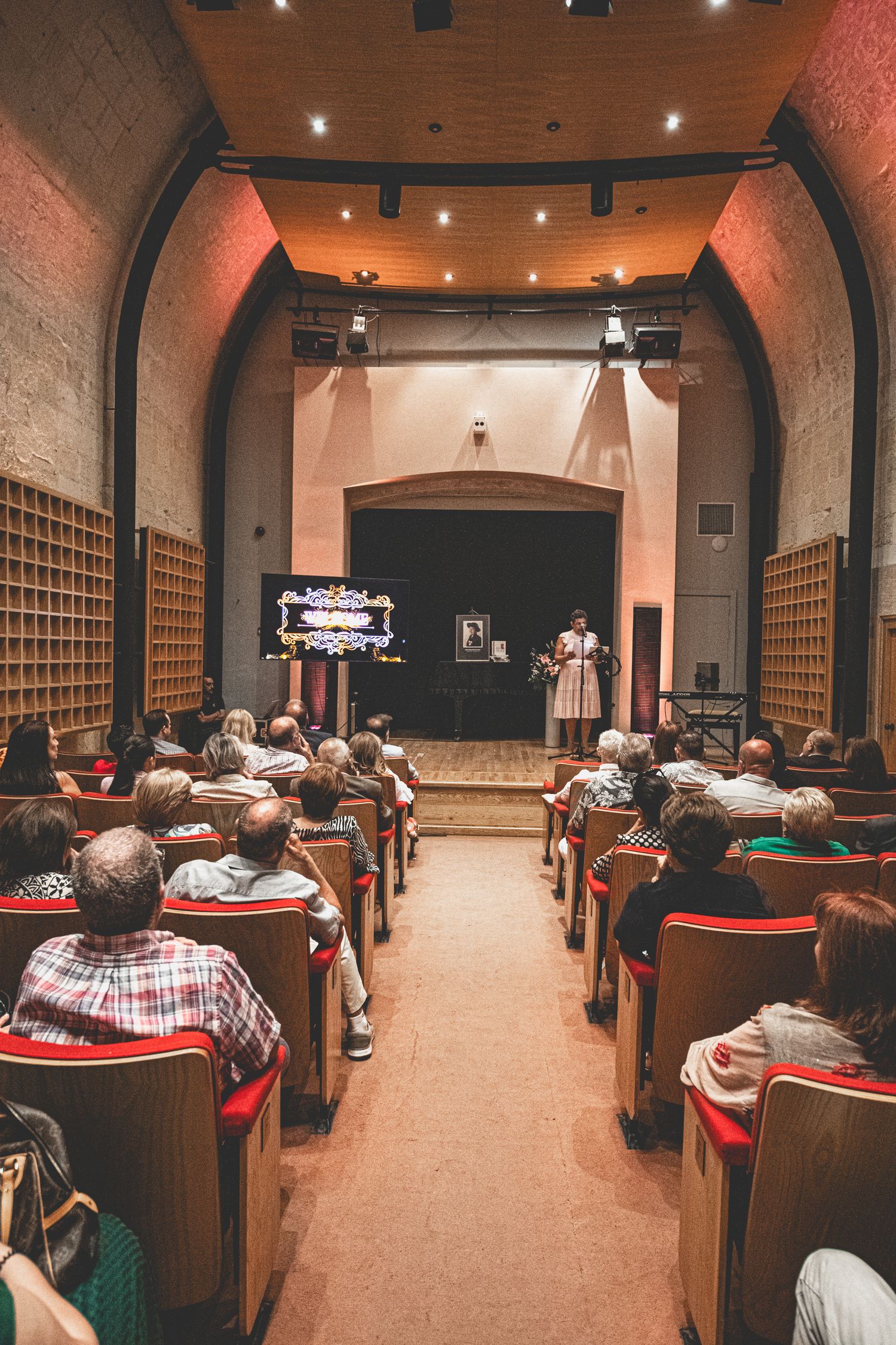 Audience seated in rows at an auditorium with arched ceiling and warm lighting.