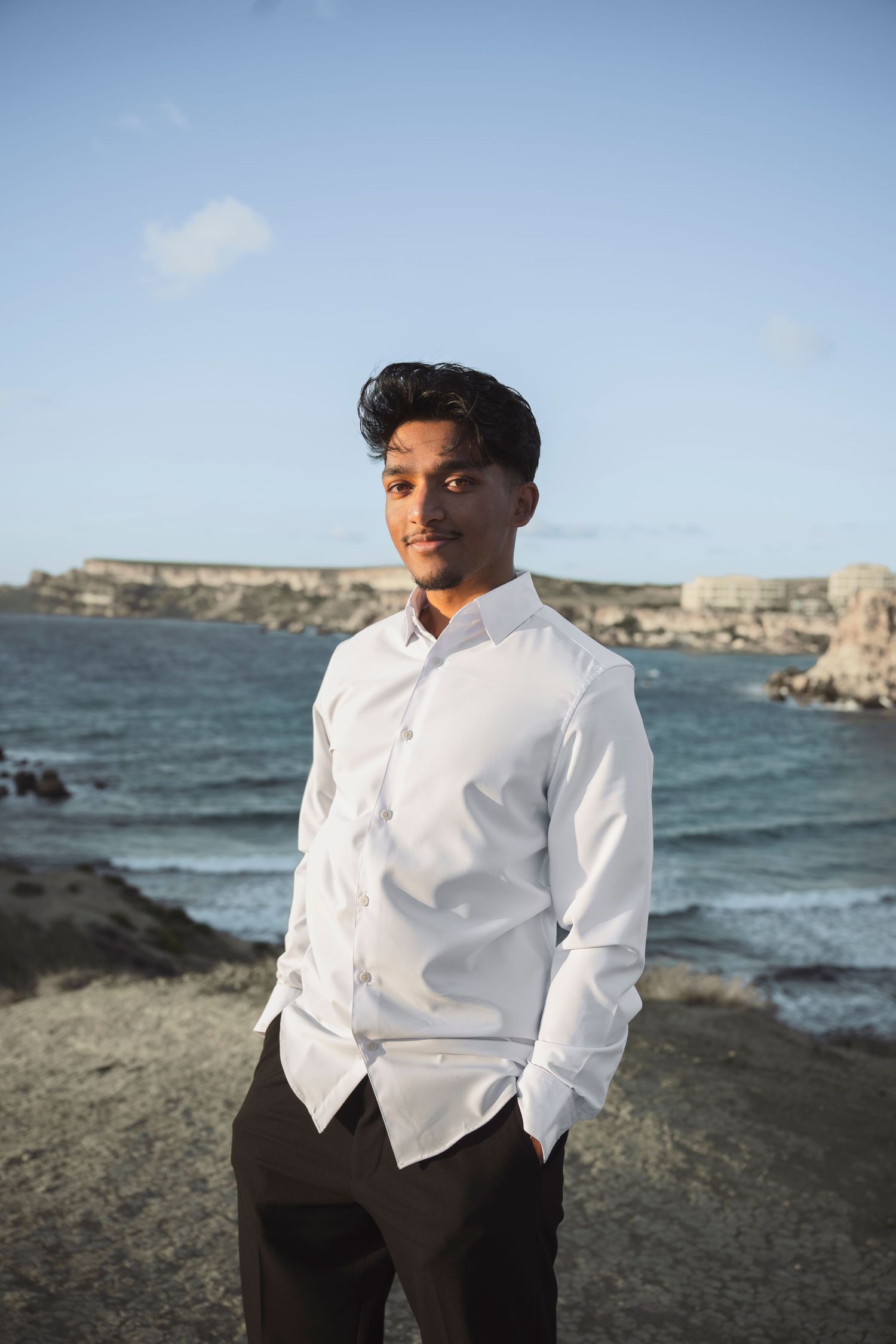 A person in a white button-down shirt poses against a scenic coastal backdrop with rocky cliffs and blue ocean waters.