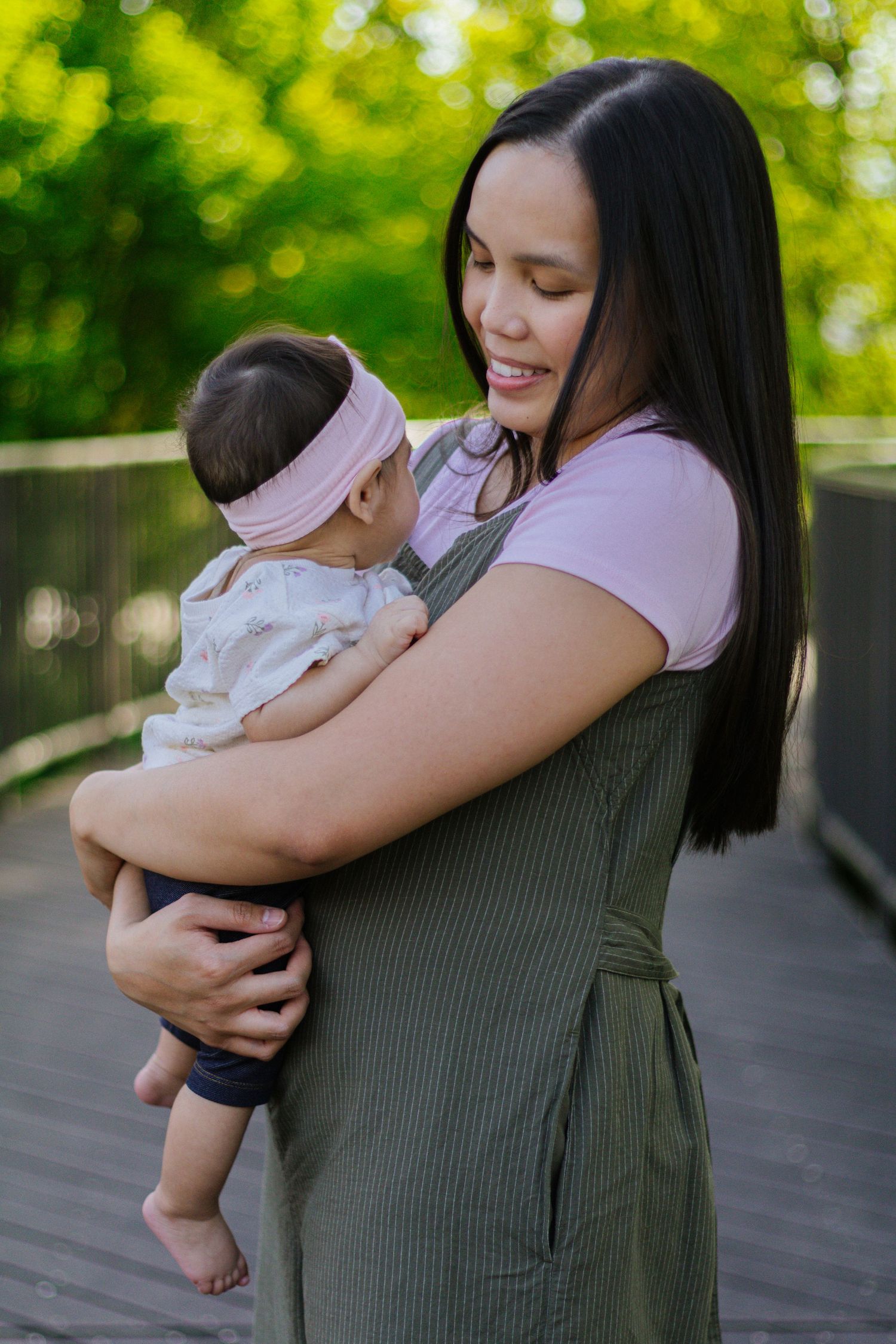 A smiling mother with long dark hair holds her baby girl outdoors on a wooden bridge surrounded by lush green trees.
