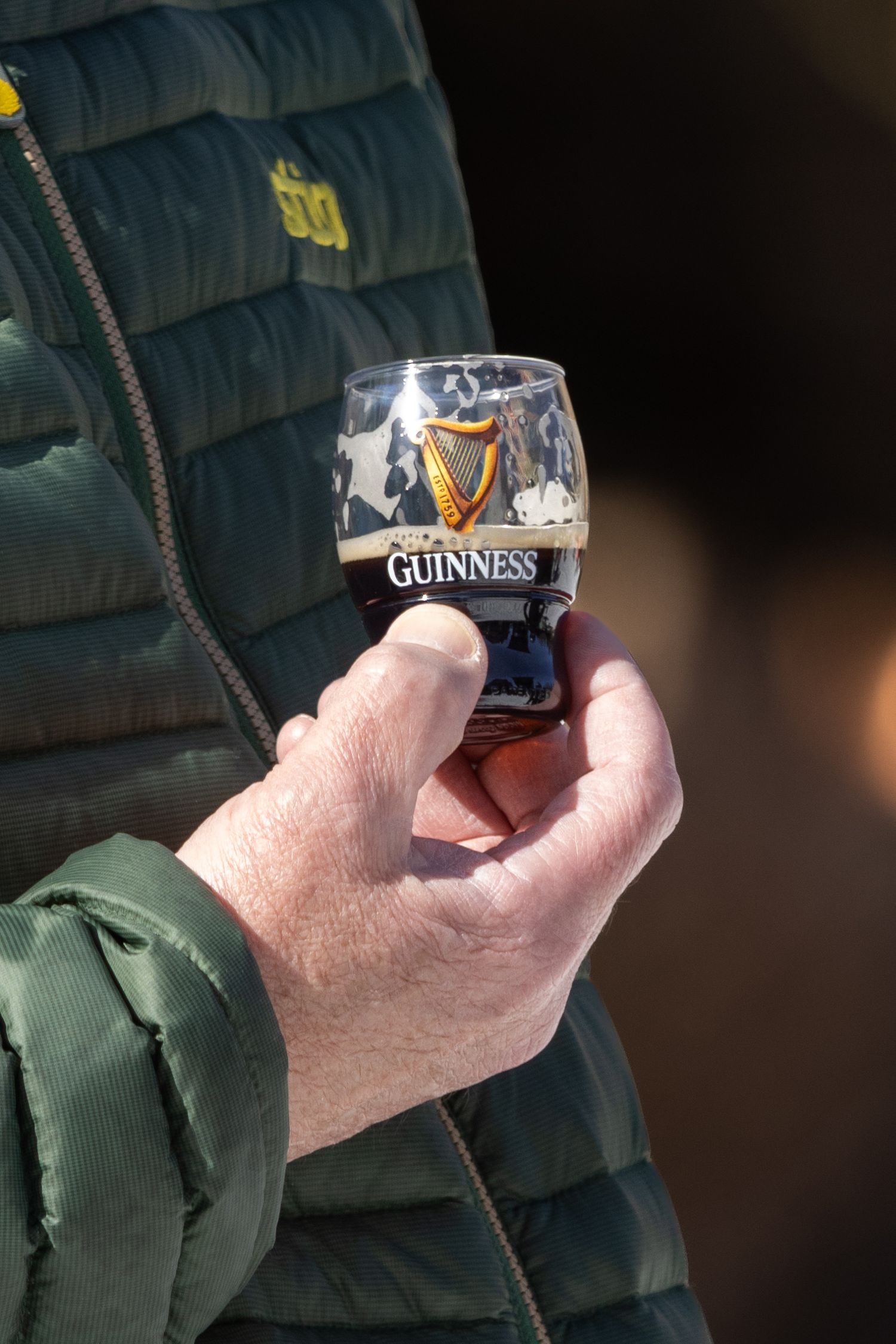 Hand holding a small Guinness beer glass outdoors, wearing a green quilted vest jacket.