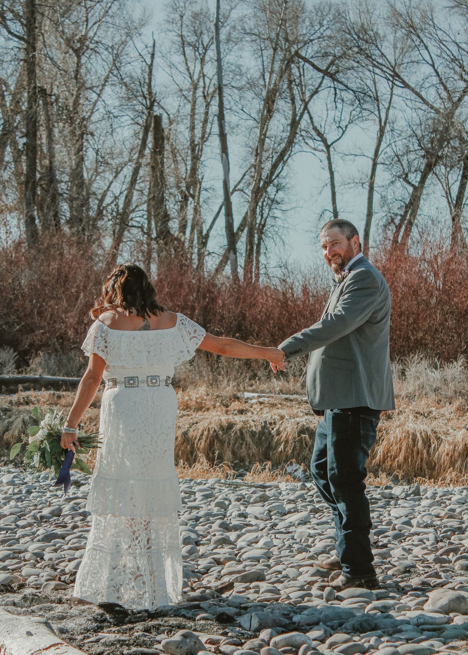 A couple shares tender moments on a rocky shore with bare trees and autumn foliage in the background.