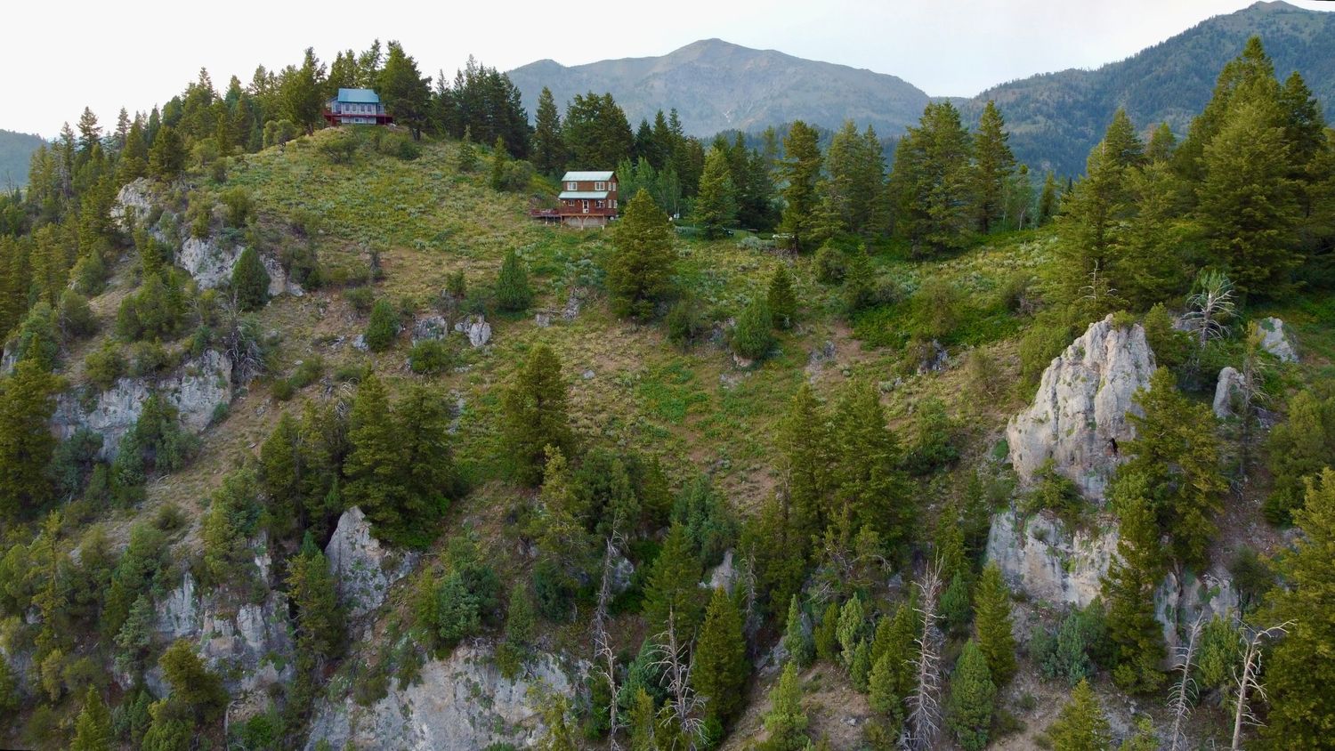 Mountain cabin perched on rocky cliffs surrounded by evergreen forest and alpine scenery.