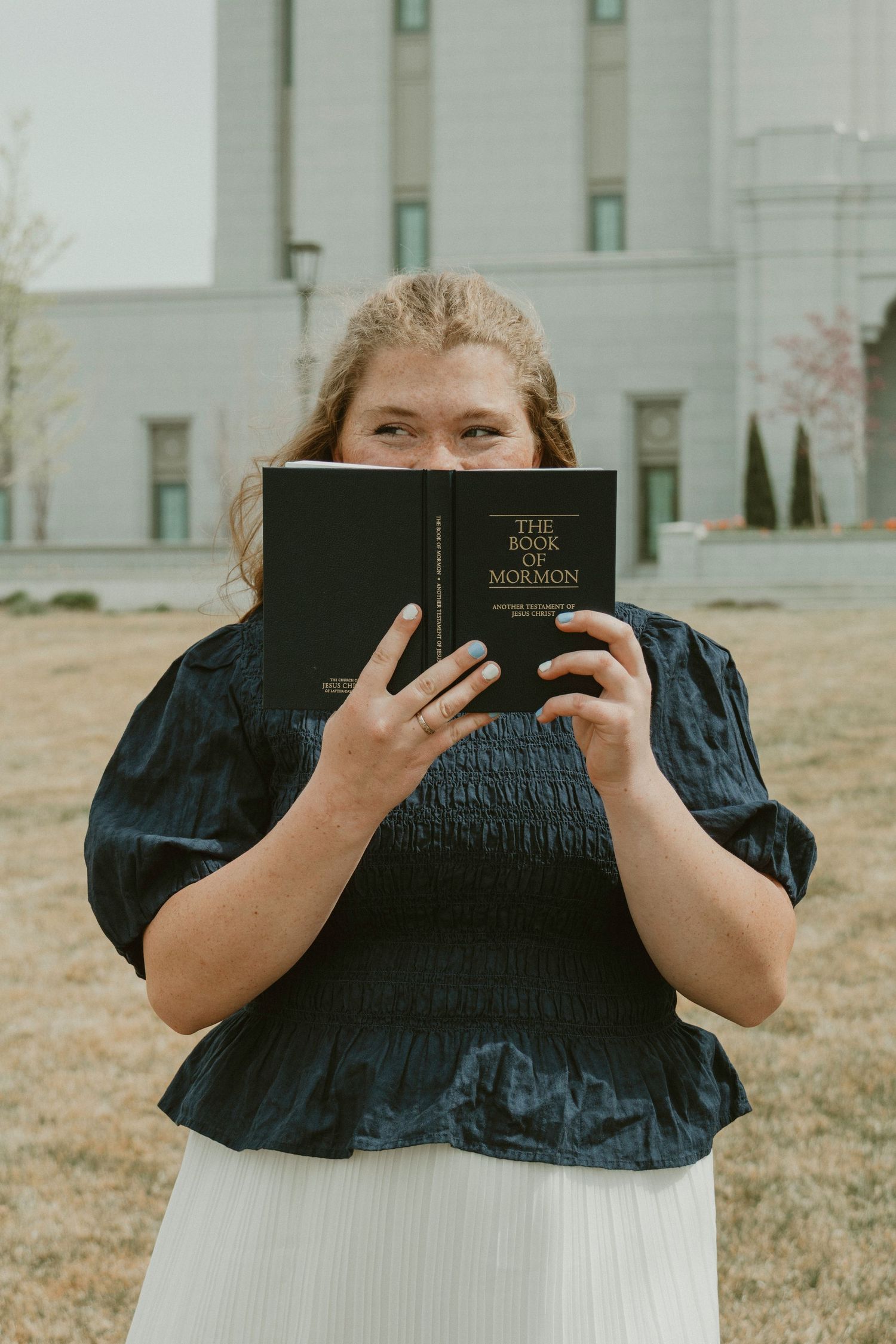 Woman holding Book of Mormon in front of face outdoors near a white building with trees in background.