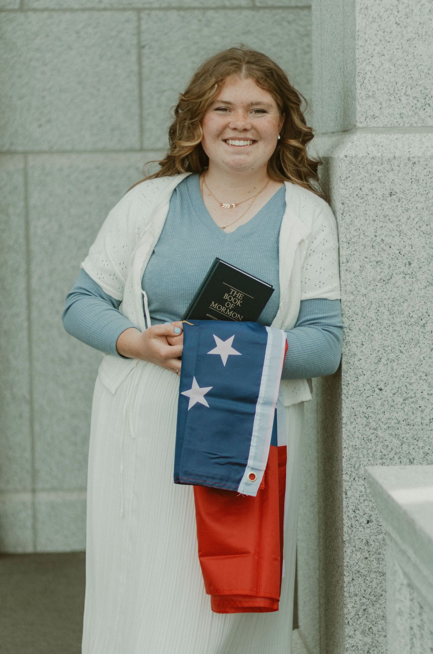 Smiling young woman holding Book of Mormon and Chilean flag, posing outdoors near stone columns.
