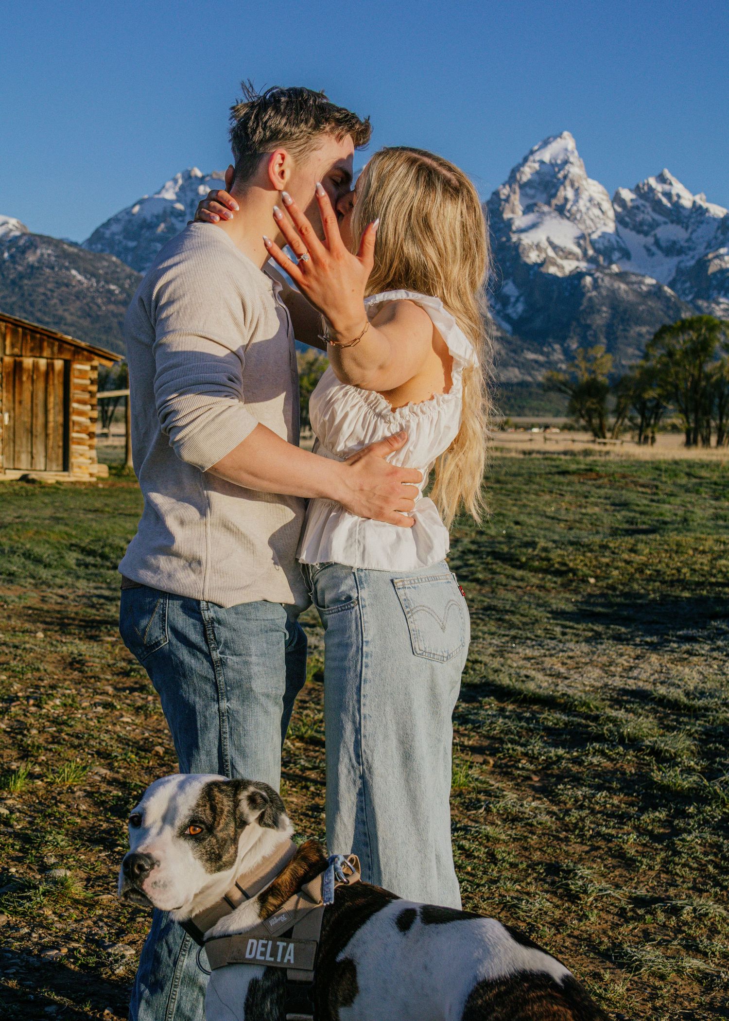 Couple kissing during engagement photo session with Grand Teton mountains backdrop and dog named Delta in foreground.