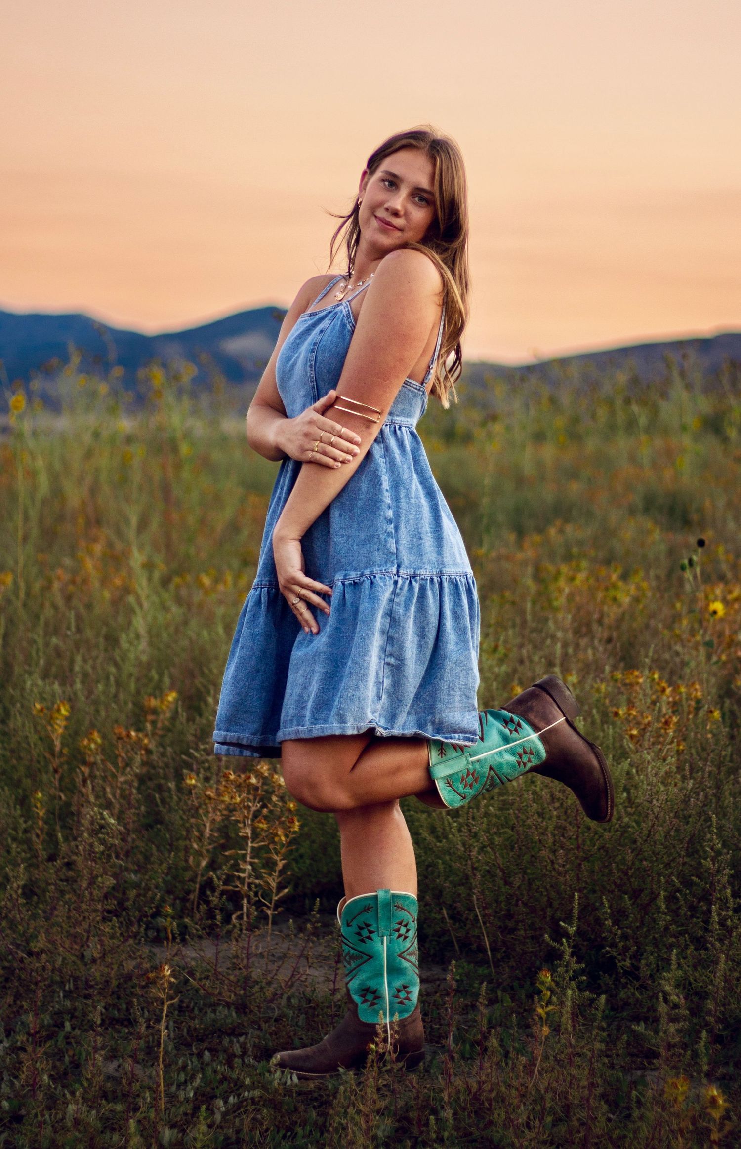Person in blue denim dress and turquoise boots poses playfully in a wildflower field at sunset.