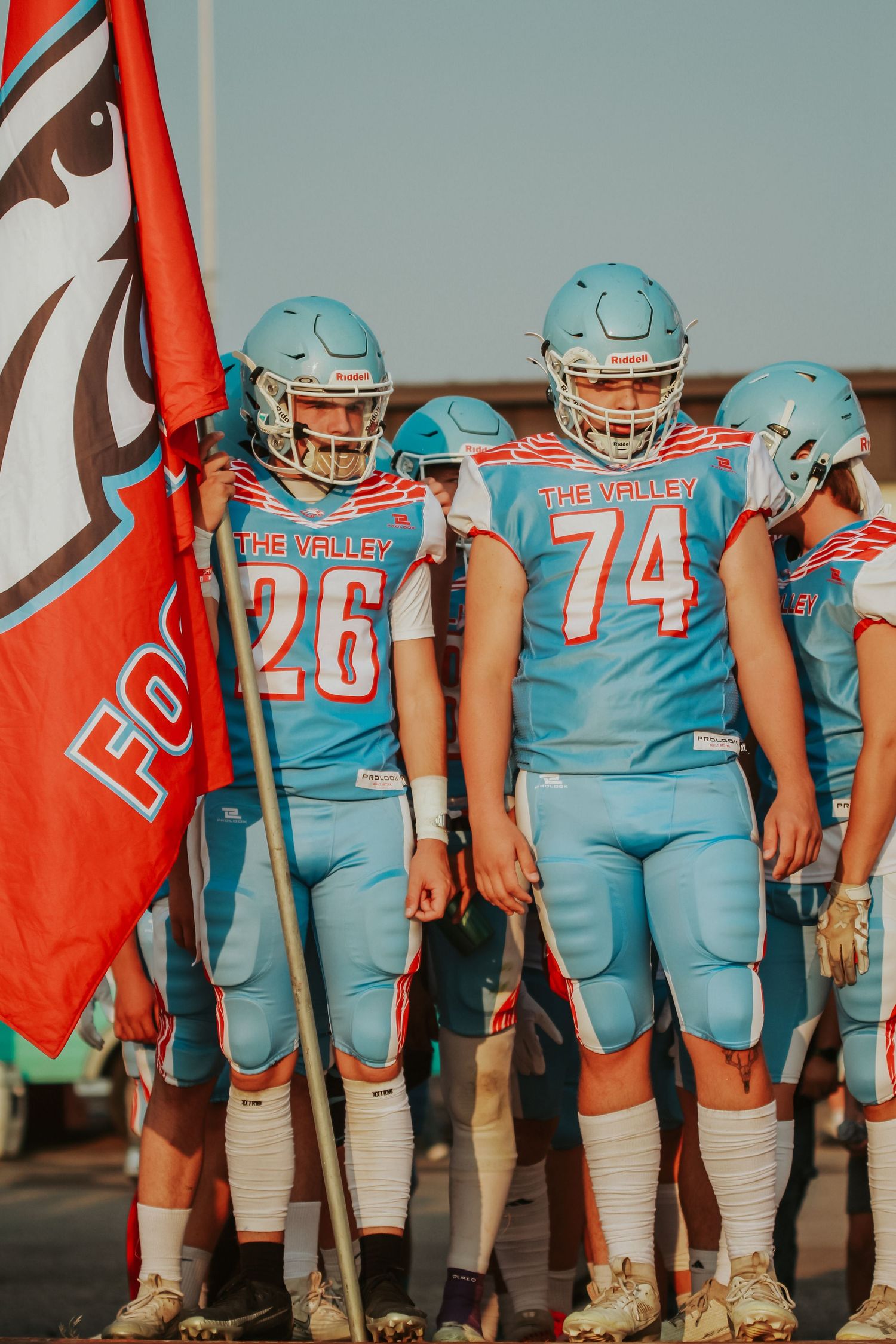 Football team in light blue uniforms stands in line formation against an orange background.