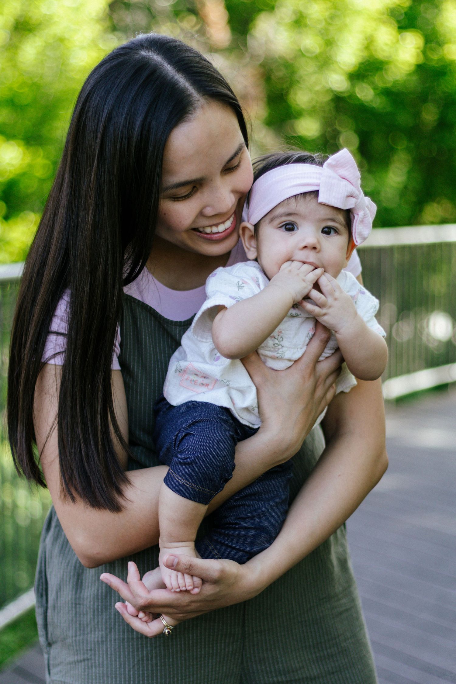 Smiling mother holding her baby girl wearing a pink bow headband outdoors in a lush green park setting.