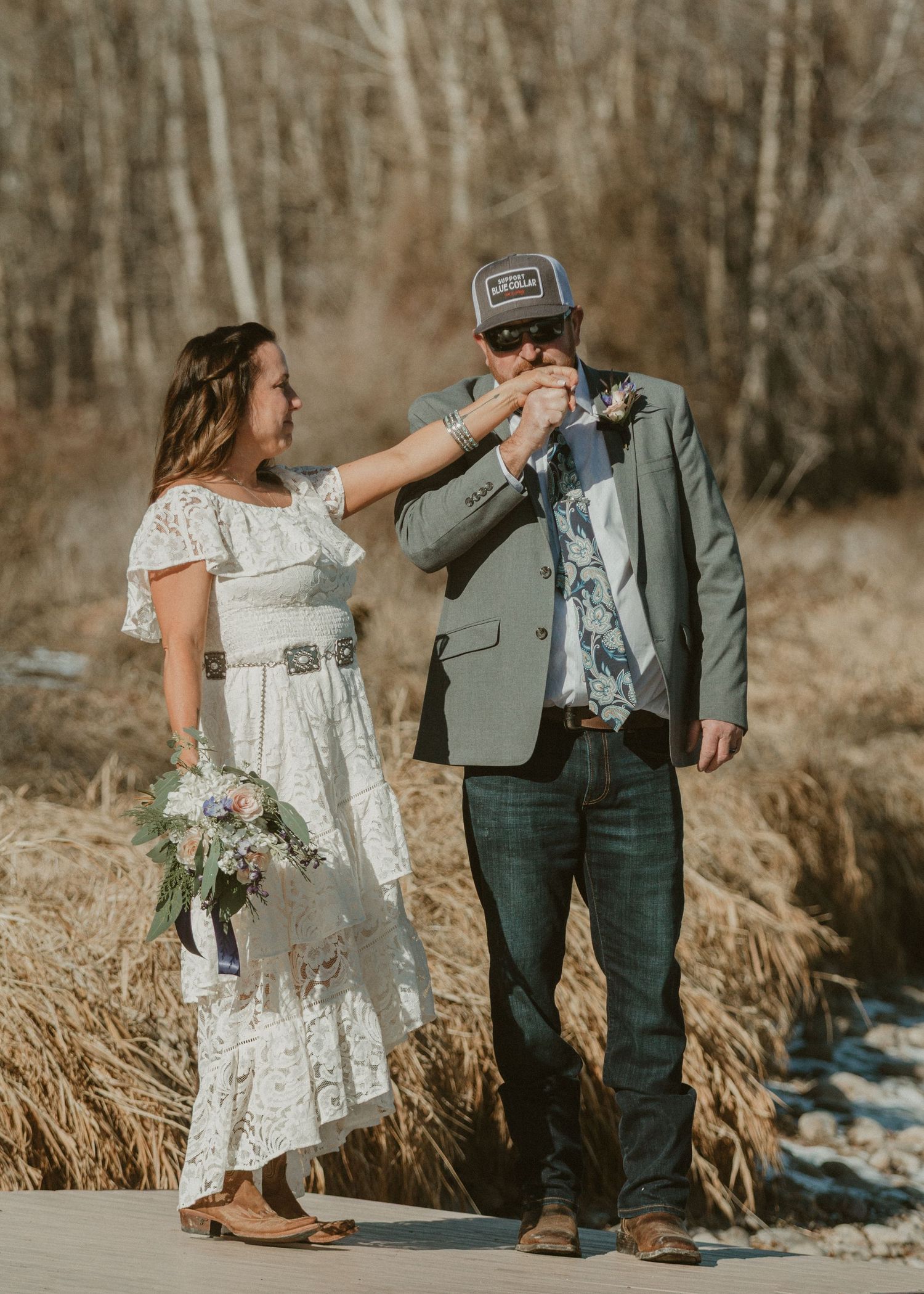 A couple in wedding attire dances together in a rustic outdoor setting with tall grass and trees in winter.