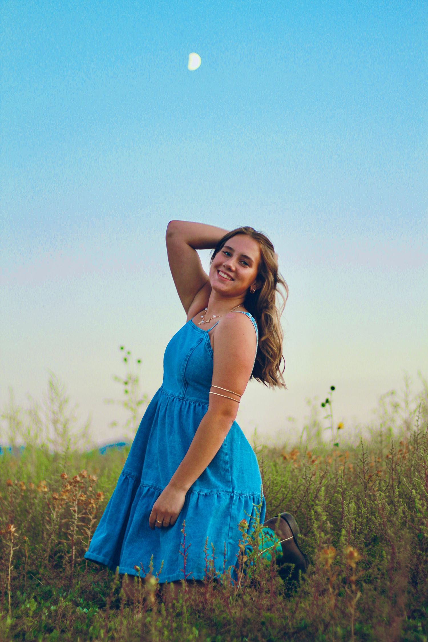 Person in blue dress poses against sunset sky with visible moon in background.