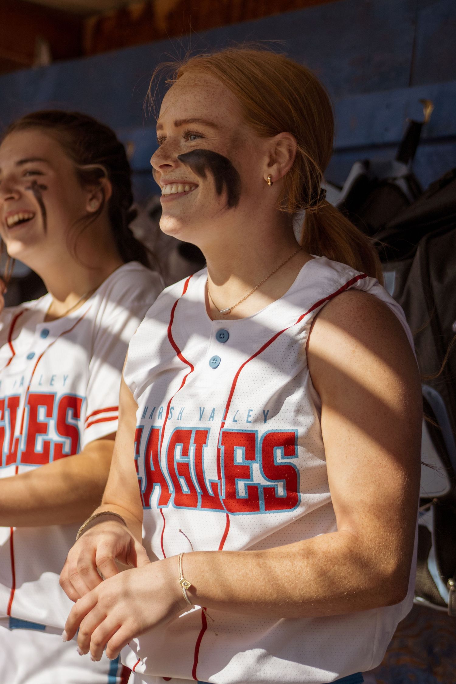 Softball players in white jerseys with red trim sitting in the dugout during a game.