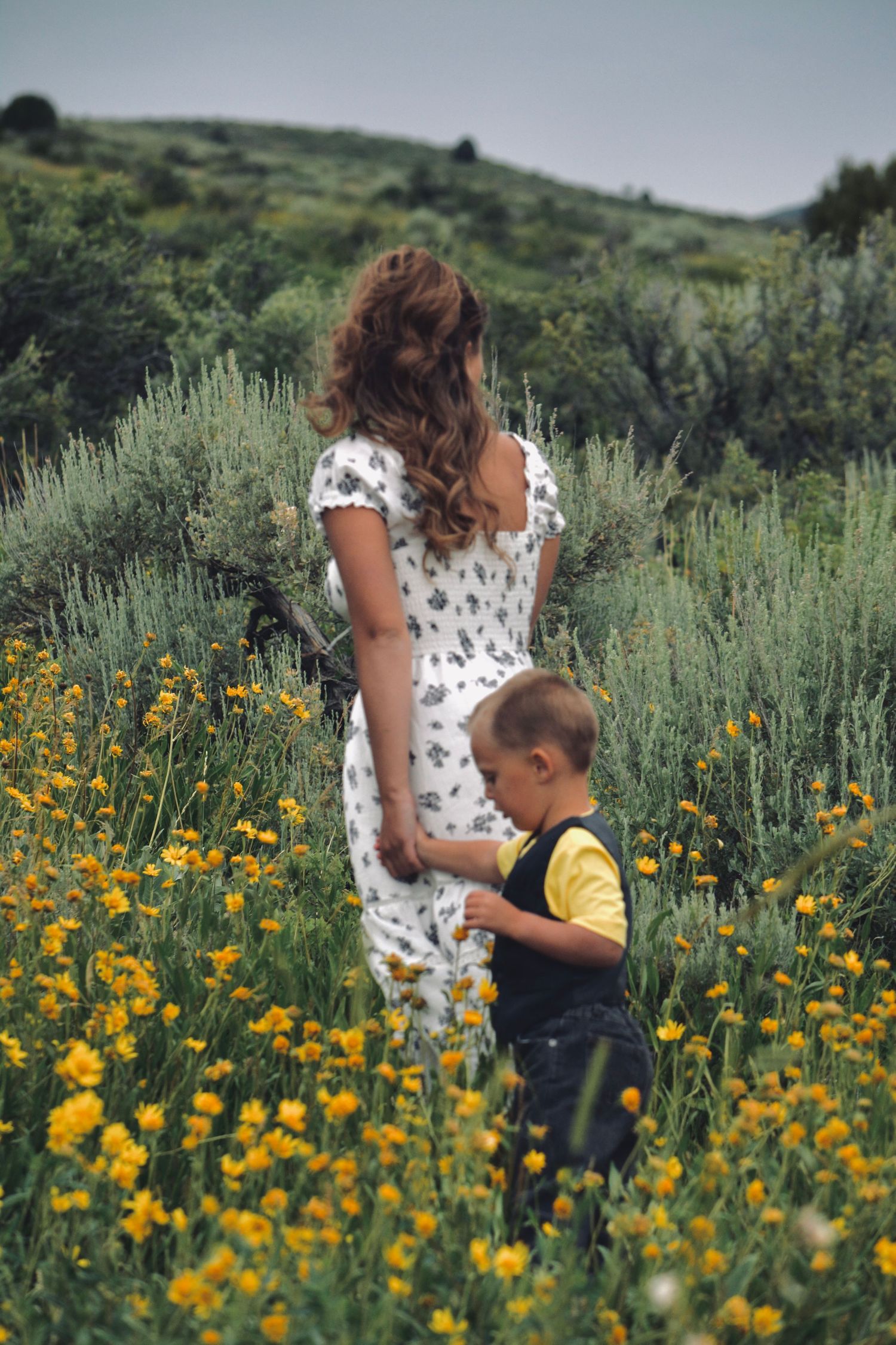 A floral dress-clad figure and child explore a wildflower meadow filled with yellow blooms on a hillside.