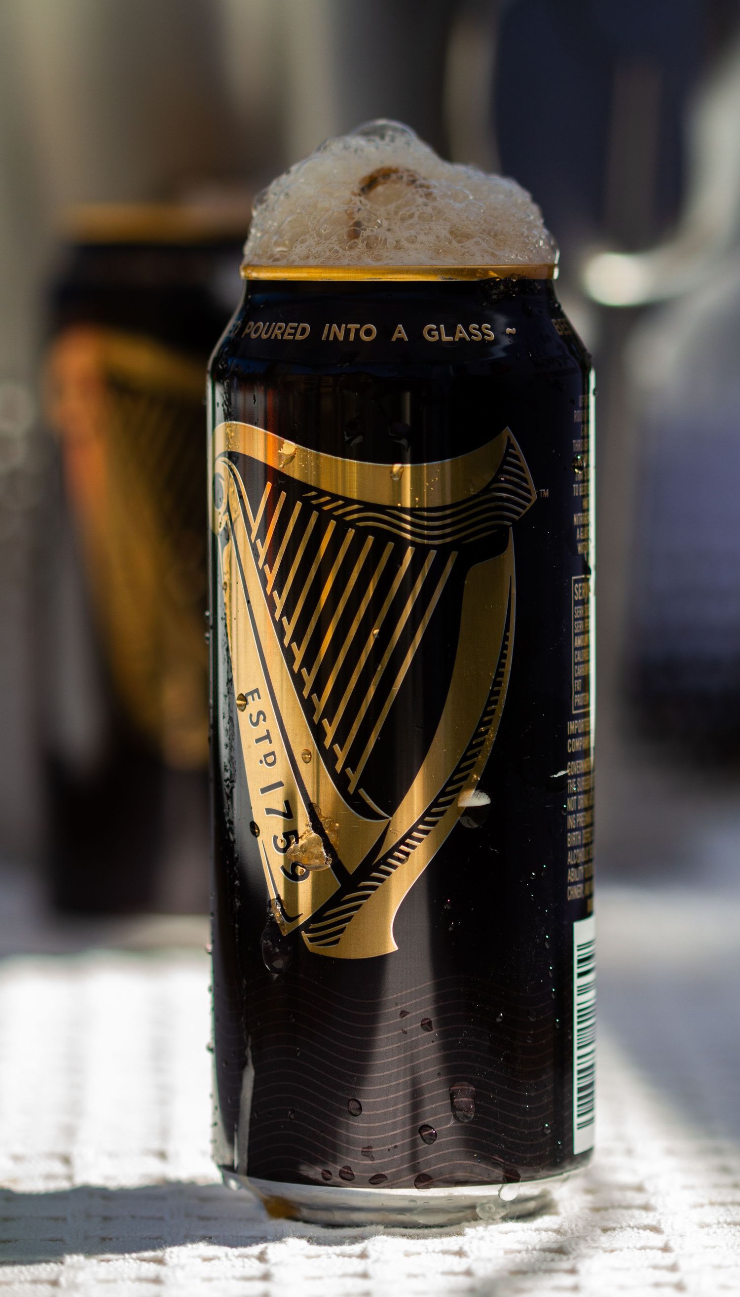 A Guinness stout beer can with a golden harp logo, overflowing with foam, sitting on a white surface.