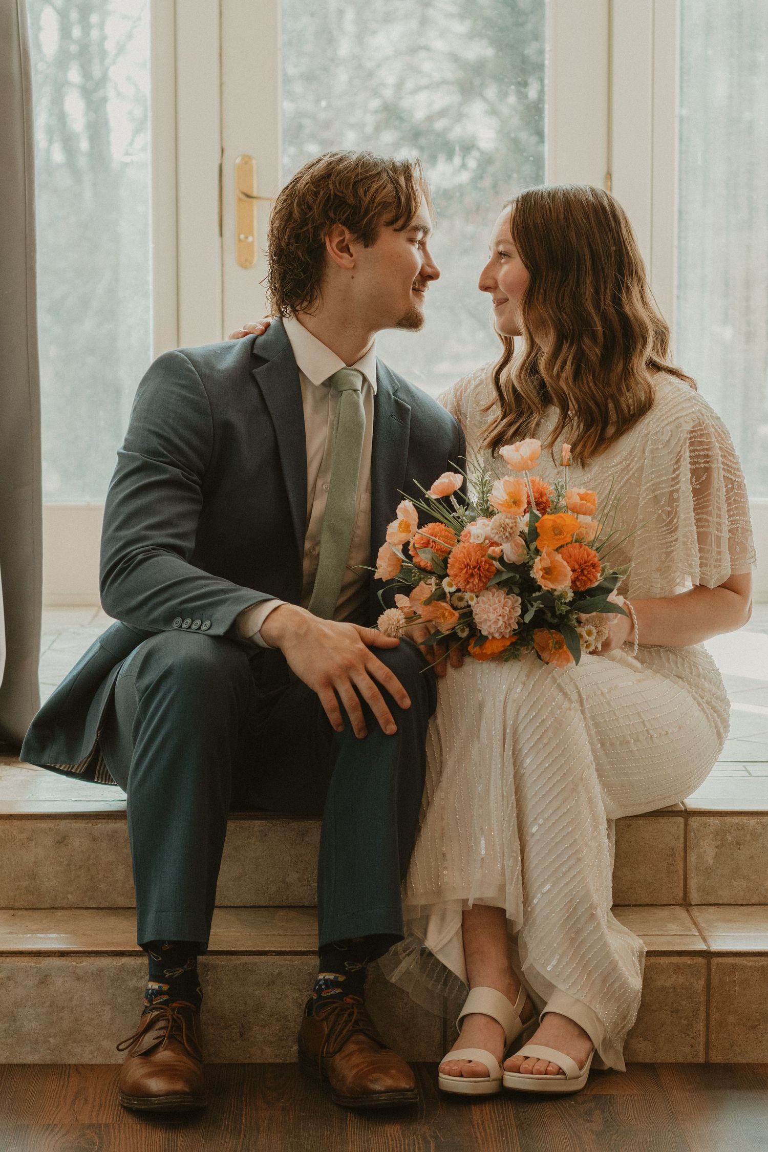 A couple in wedding attire share a joyful moment on a window ledge while holding a peach and coral floral bouquet.