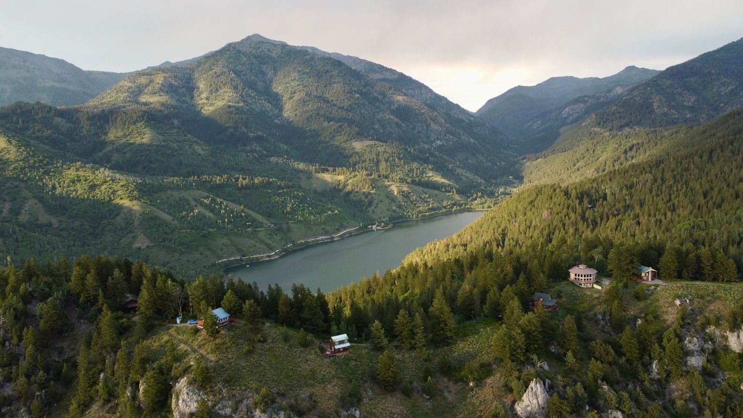 Sunset view of mountain valley with lake nestled between forested peaks and scattered buildings.