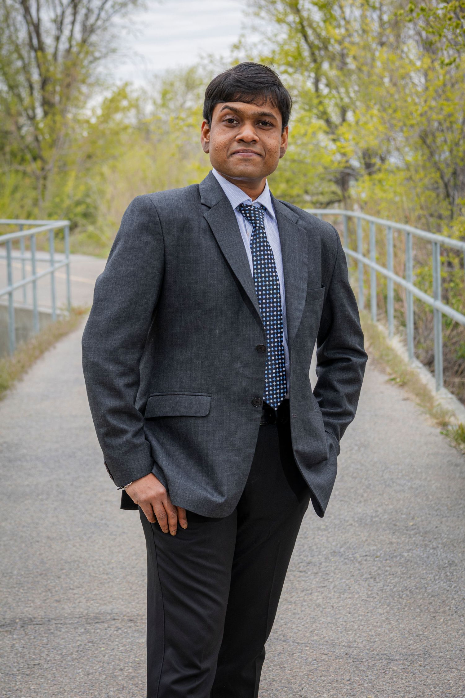 Man in gray suit and blue tie stands on outdoor bridge path surrounded by trees.