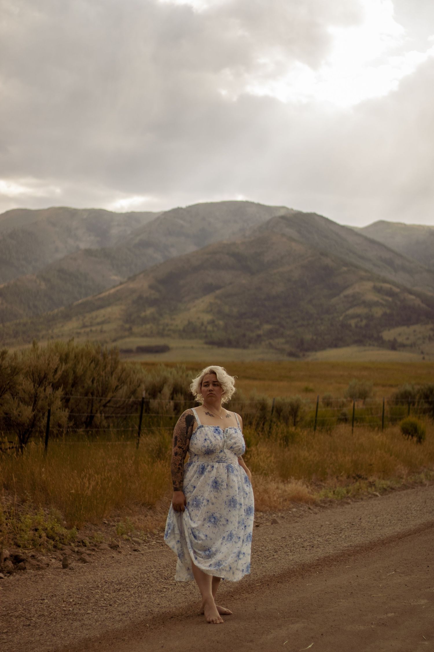 Person in floral dress walks barefoot along mountain path during cloudy sunset.