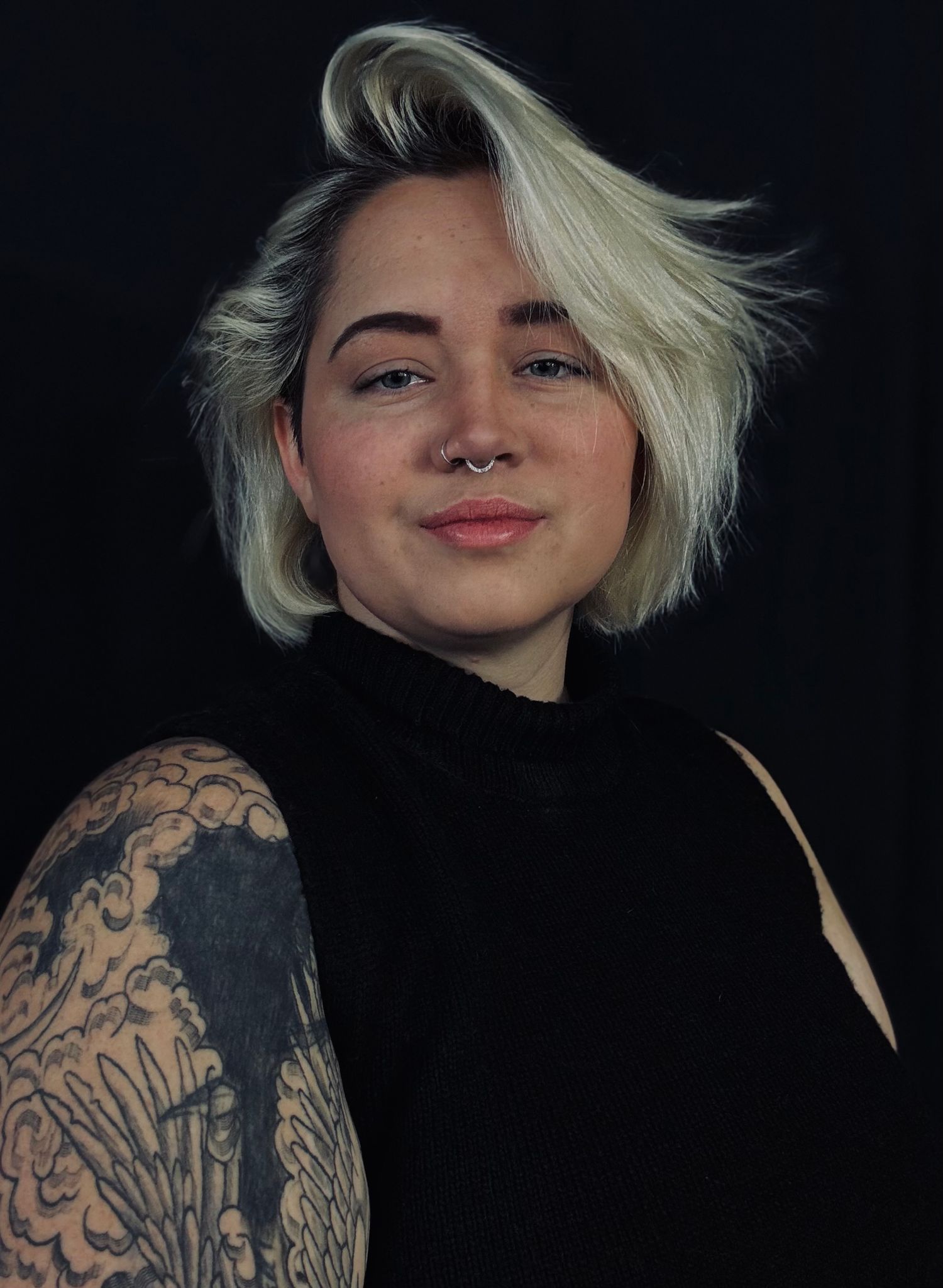 Studio portrait showing tattooed arm and black turtleneck against dark background.