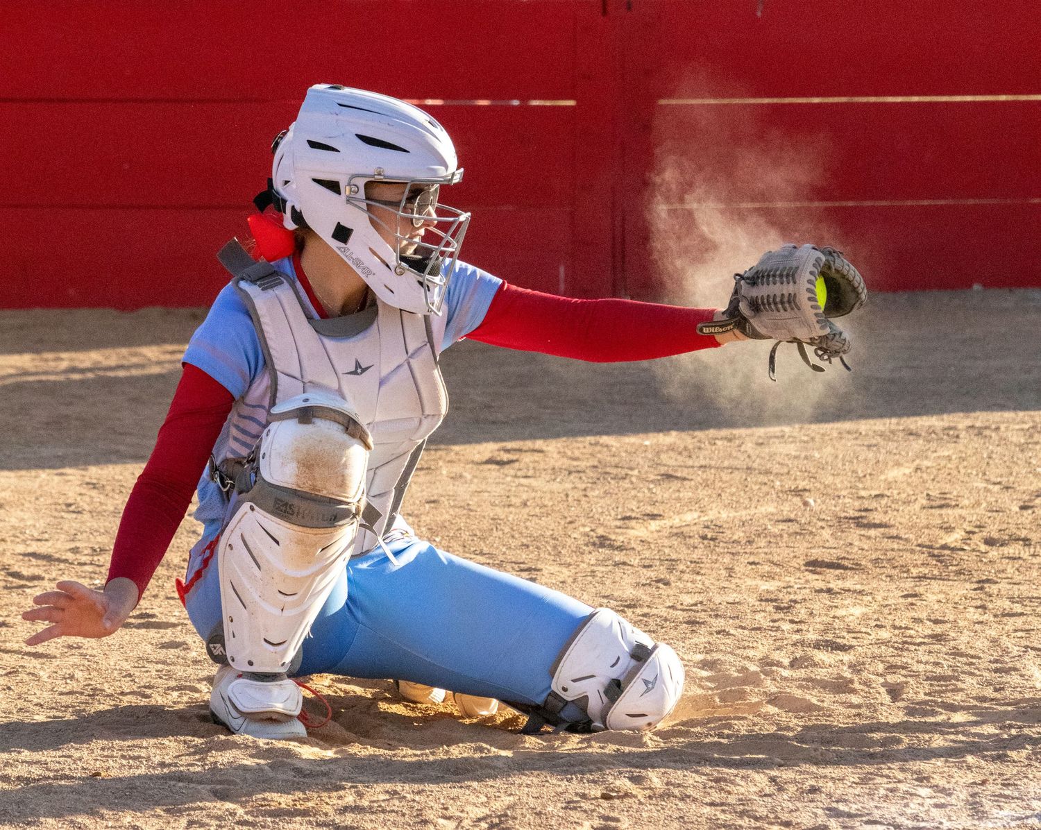 Softball catcher in white helmet and gear crouching to catch a pitch in a dusty dirt field.