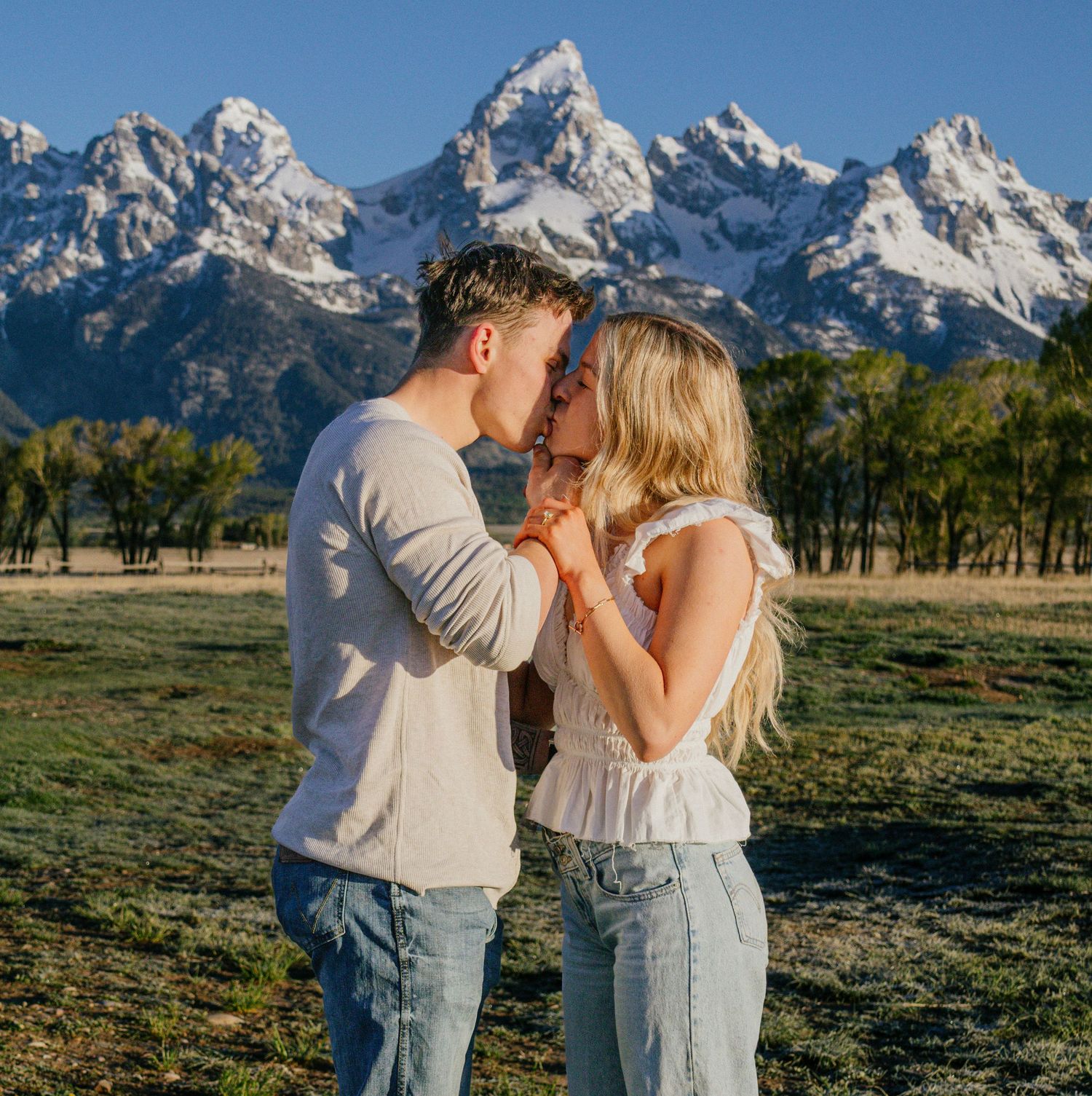 A couple shares a romantic kiss in a sunlit meadow with the snow-capped Grand Teton mountains in the background.