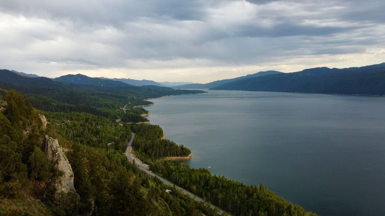 Dramatic landscape of a mountain lake with winding shoreline road and forested hills.