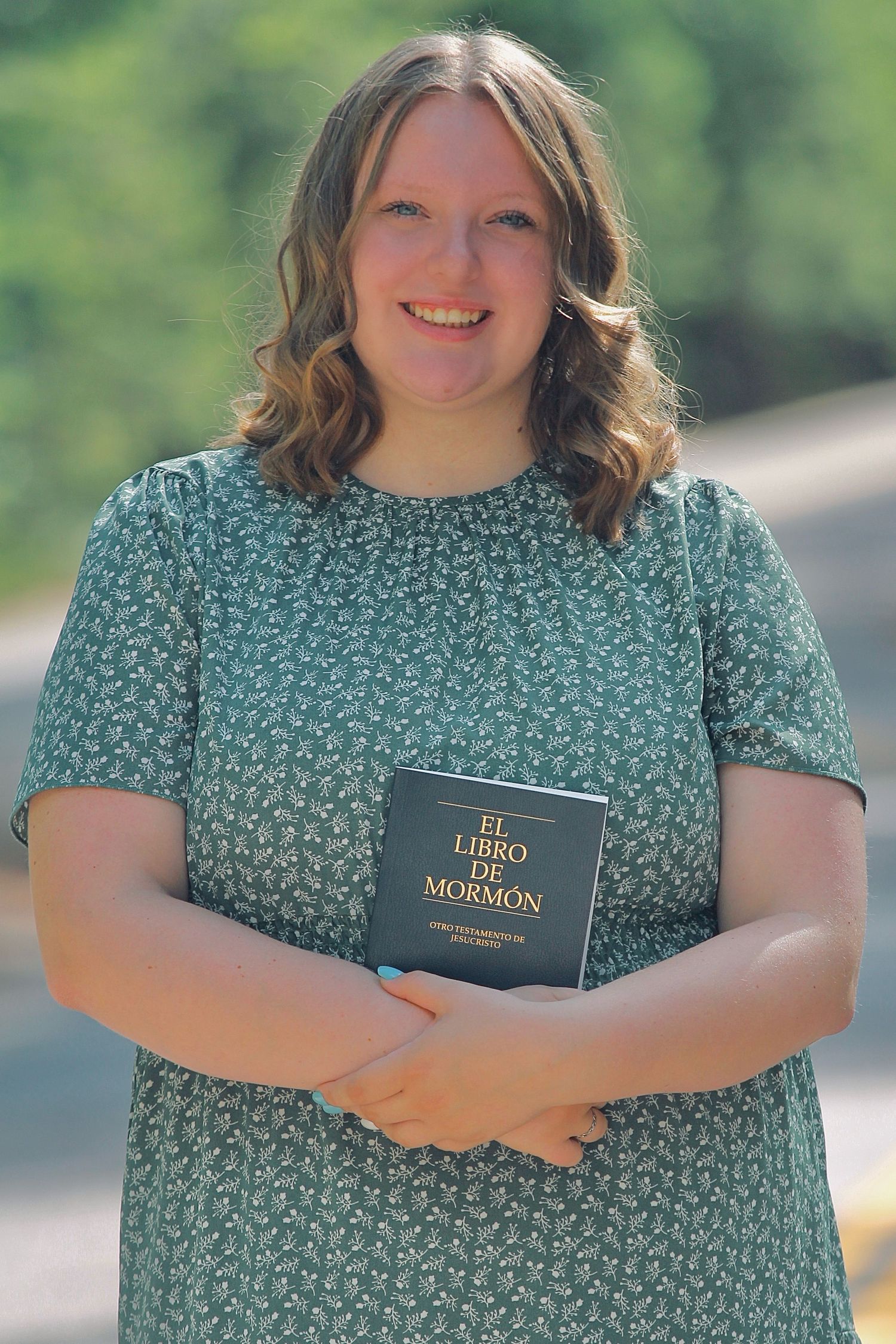 Person in a green floral dress holds a Book of Mormon while standing outdoors.