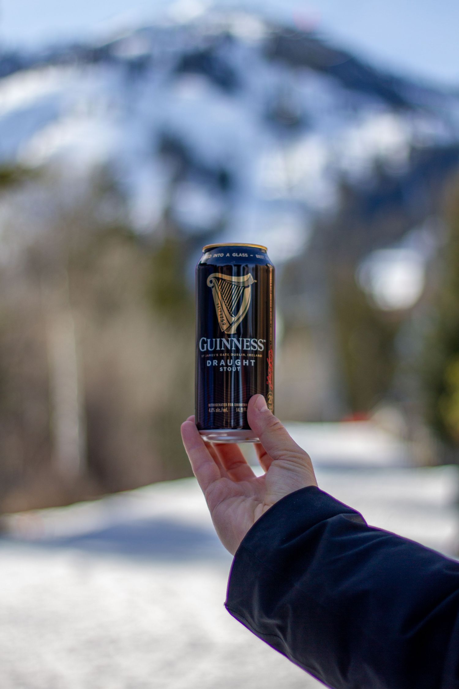 Hand holding a Guinness Draught can against a snowy mountain backdrop on a sunny winter day.