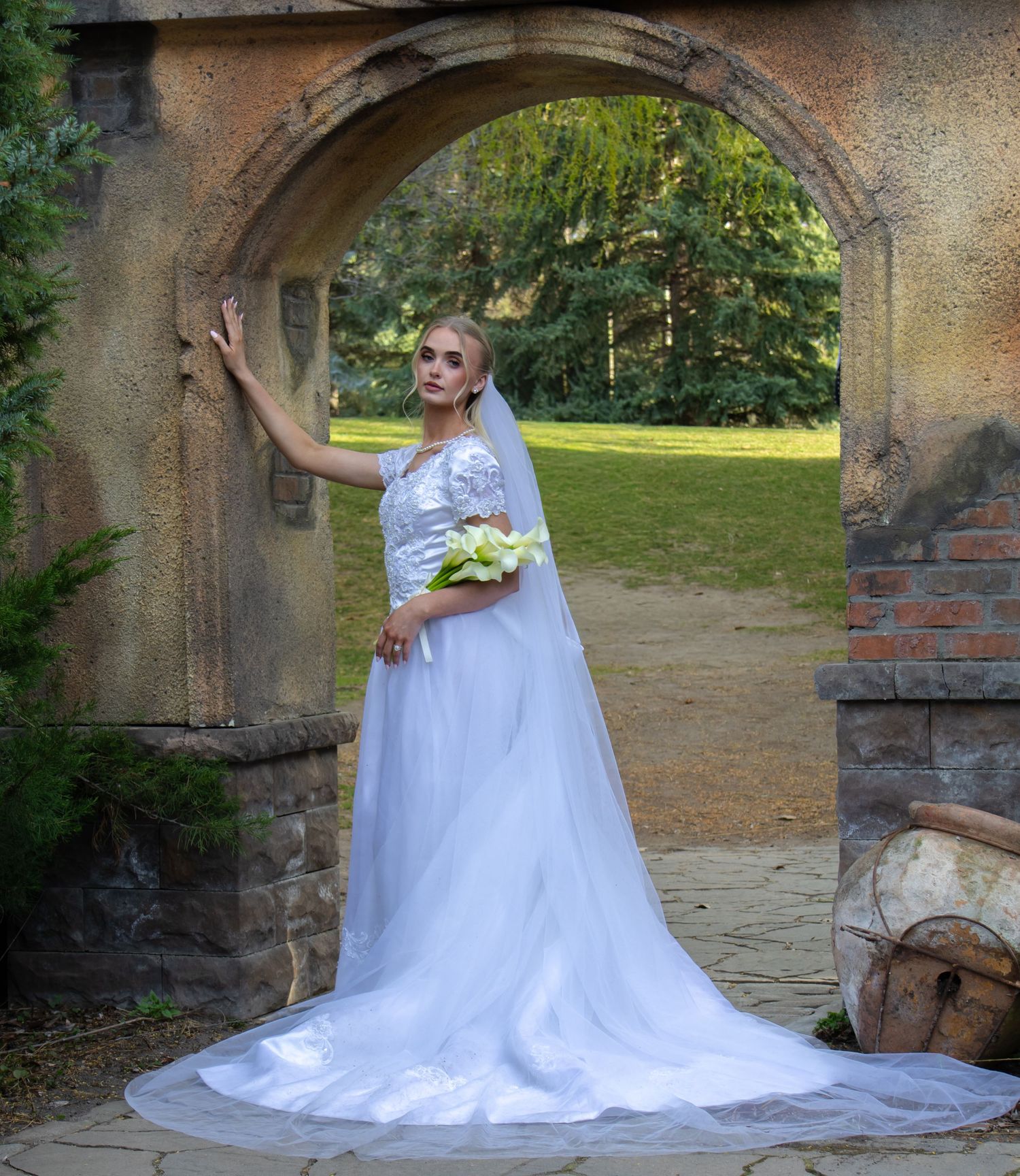 A bride in a flowing white wedding gown with a long train poses in a garden setting under stone archways.