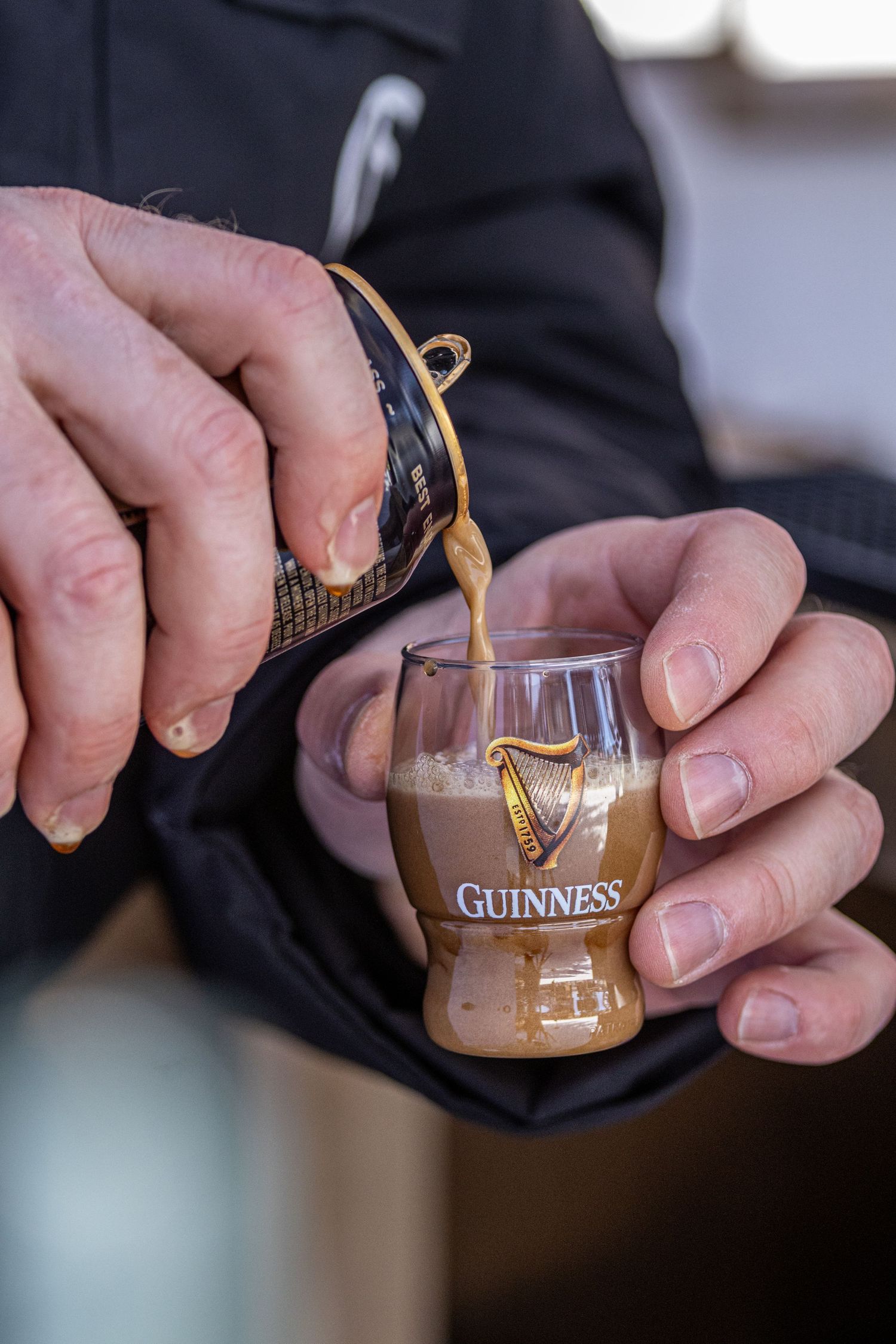 Hands pouring Guinness beer into a small branded shot glass, with creamy foam forming at the top.