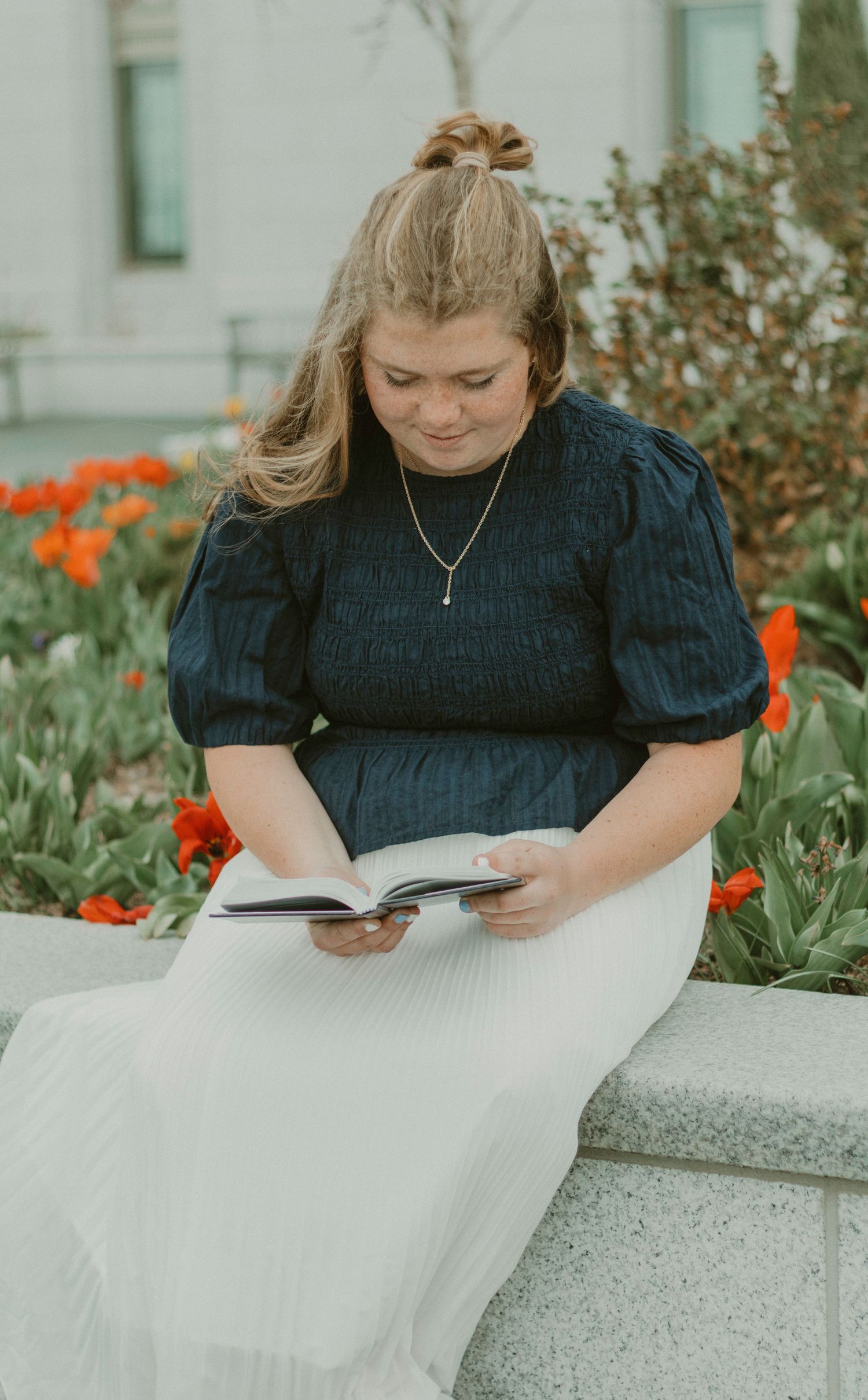 Young woman in dark top and white skirt reads a book outdoors near red tulips in a garden setting.