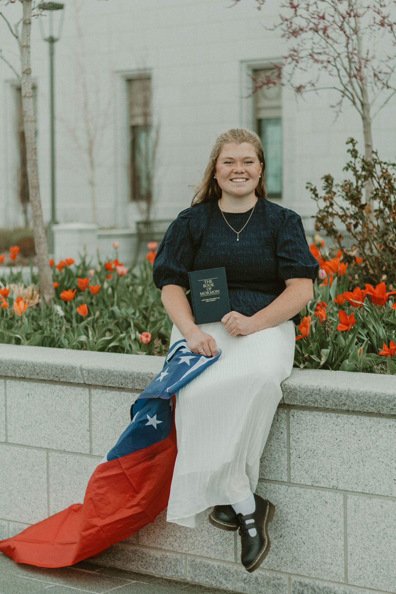 Young woman sitting outdoors holding a book and flag, smiling among orange tulips near a white building.
