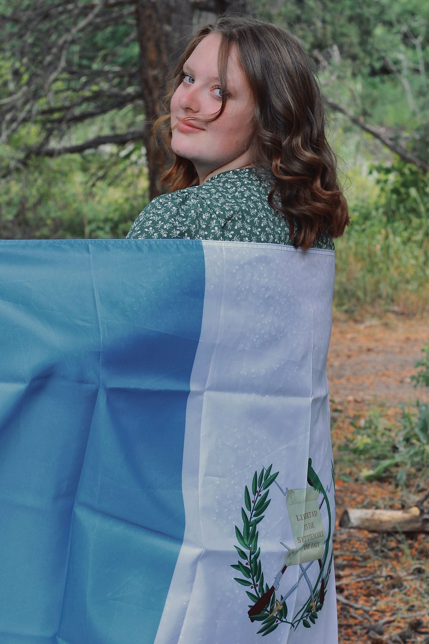 Person wrapped in Guatemalan flag poses outdoors in green dress.