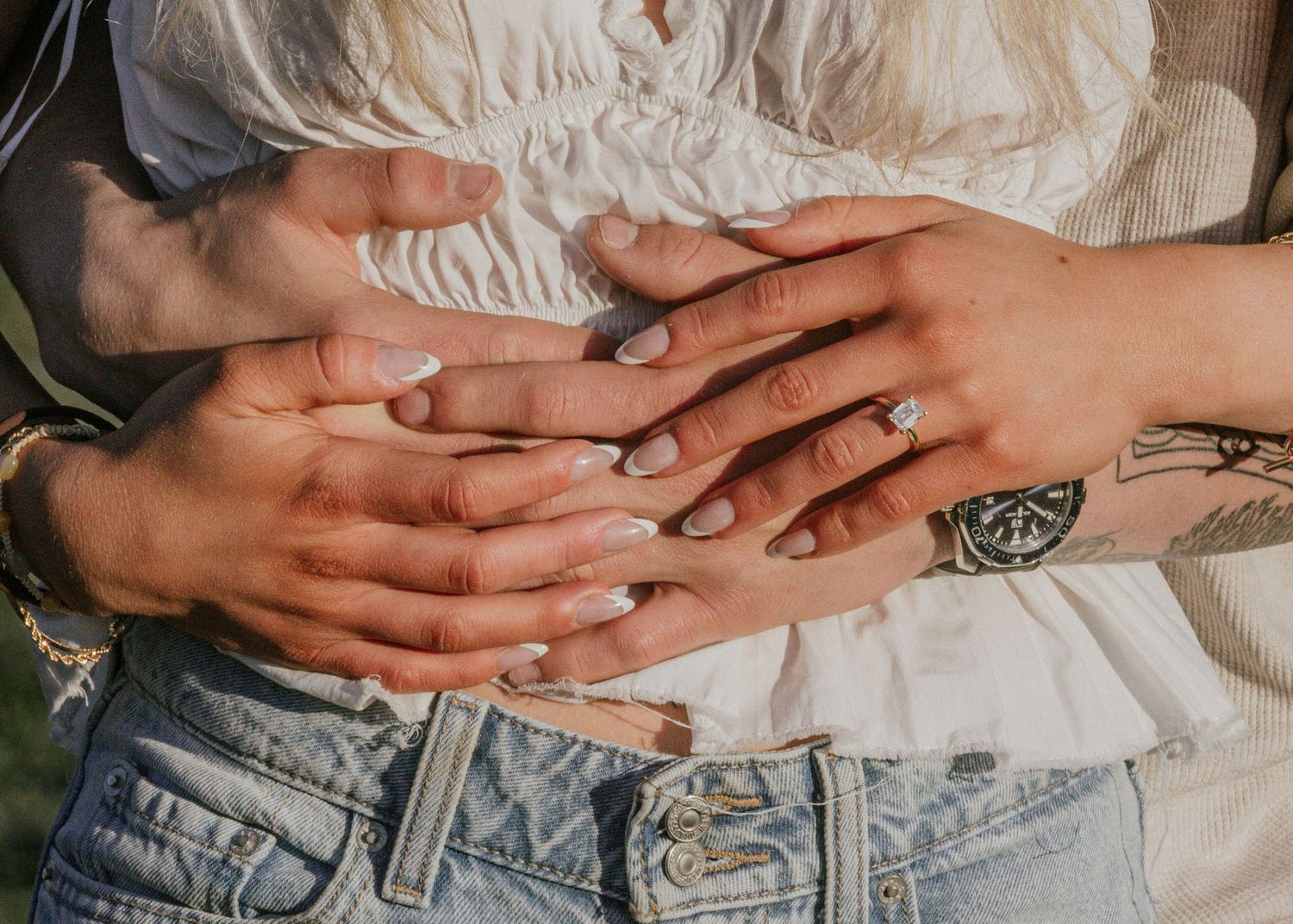 Close-up of two people holding hands showing an engagement ring, with tattoos visible on one wrist, wearing jeans.