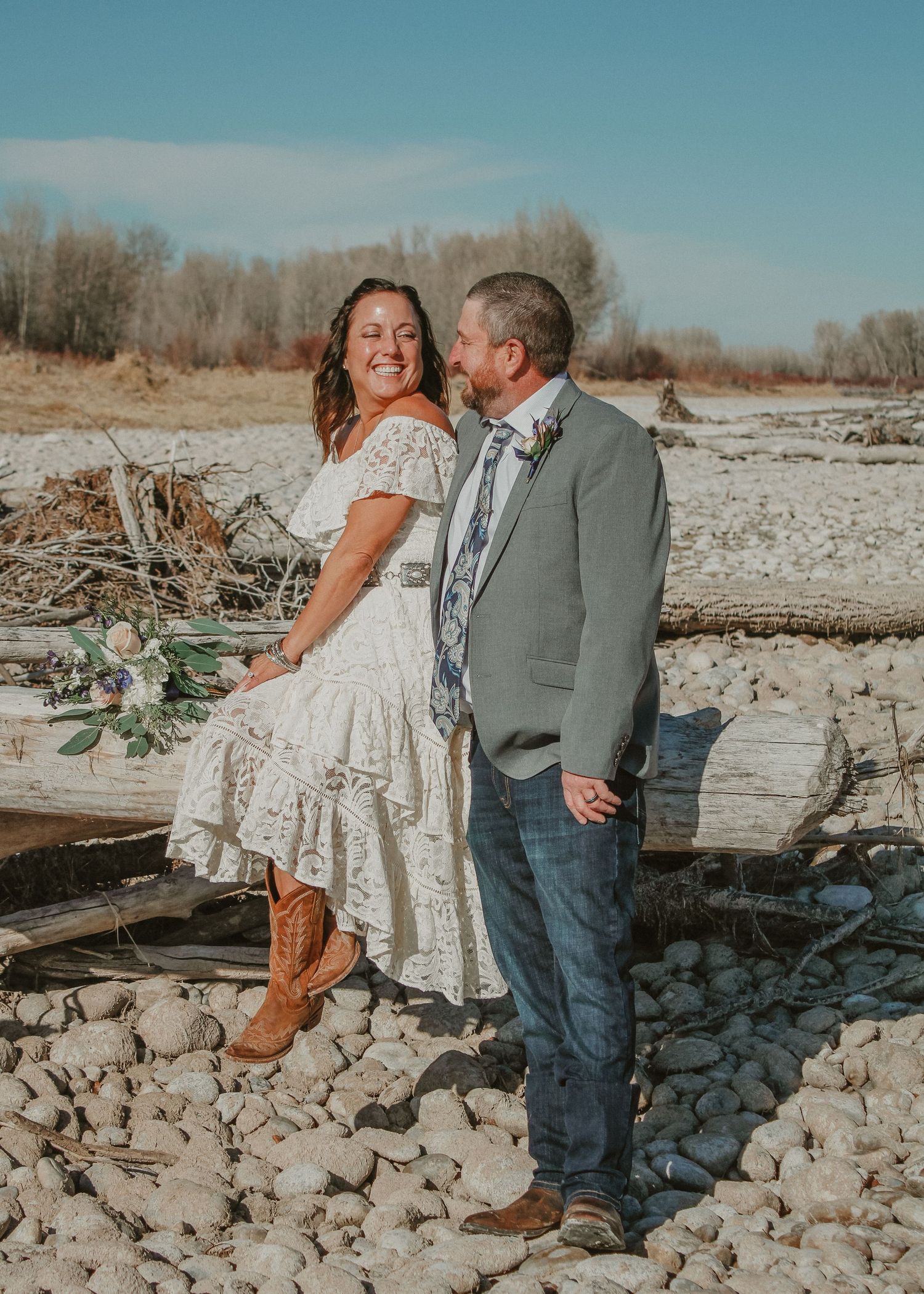 A romantic couple in western attire poses along a rocky riverbank during a winter photo session.