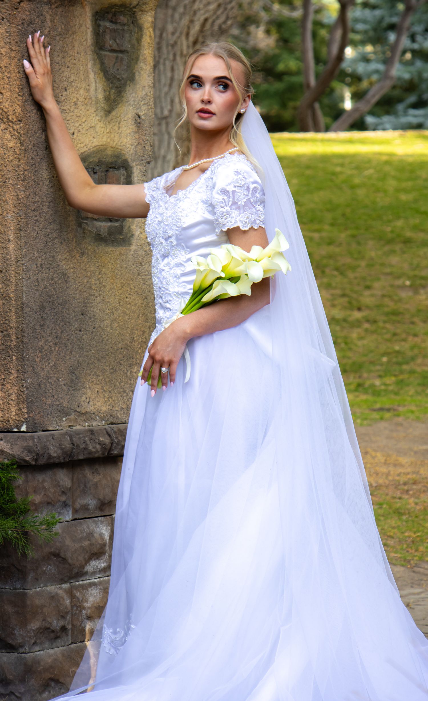 A bride in an elegant white lace wedding gown with long sleeves poses against a stone wall in an outdoor garden setting.