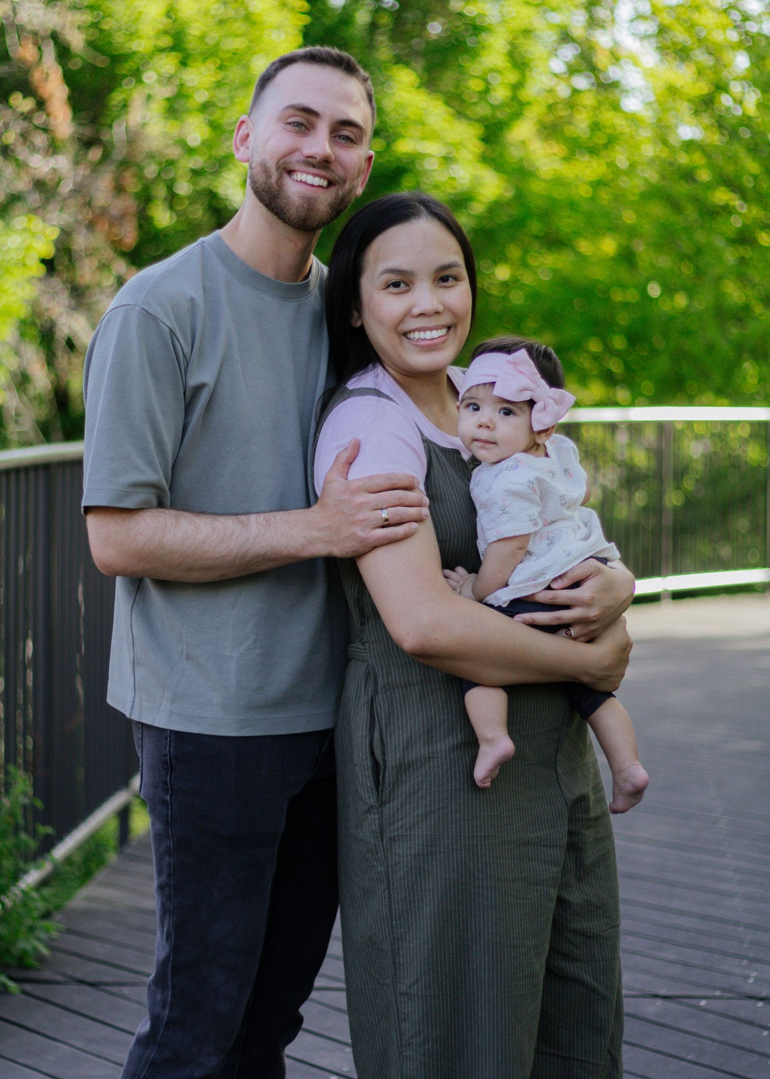 A smiling couple stands on a wooden deck outdoors, with the woman holding a baby girl wearing a pink bow headband.