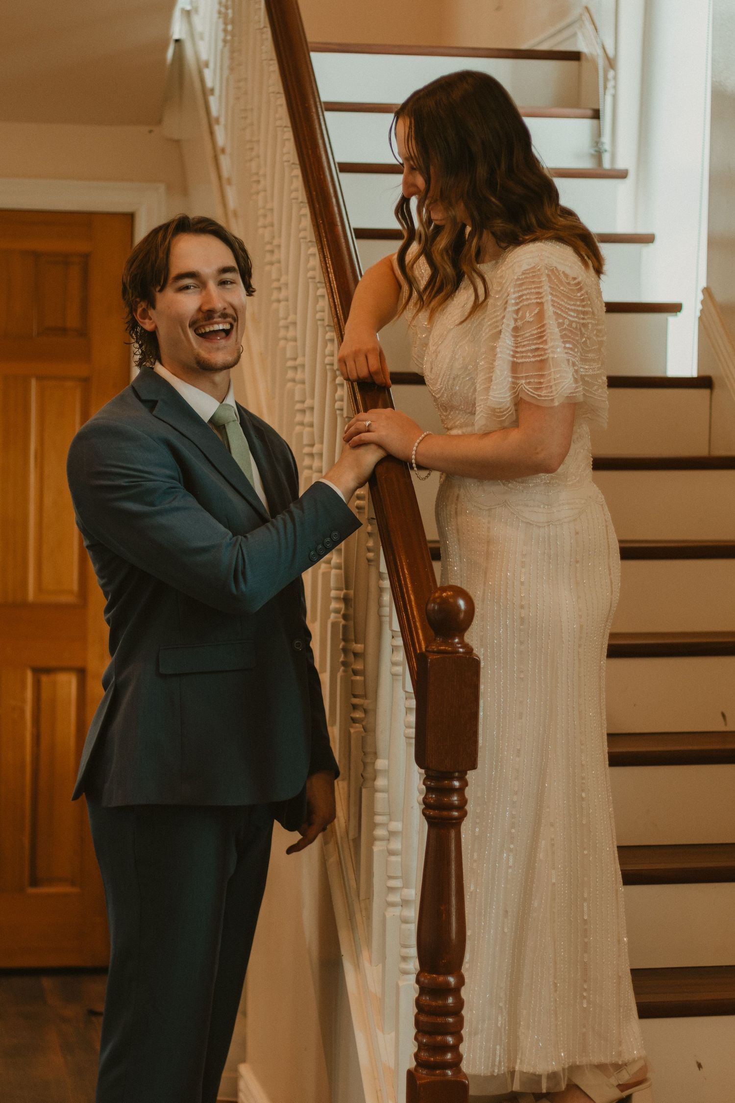 A couple shares a joyful moment on a wooden staircase, with the person in a navy suit proposing to someone in a white dress.