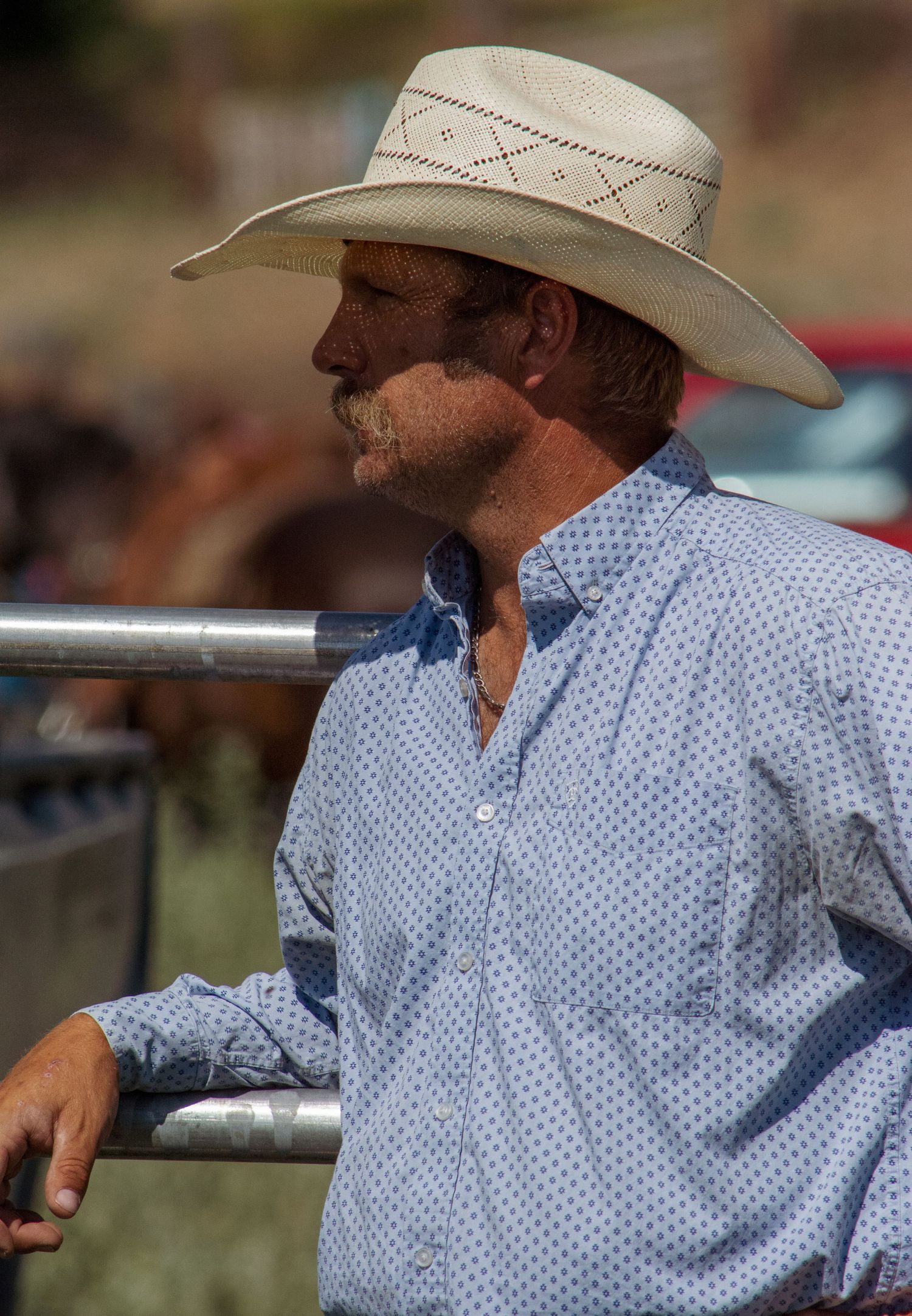 Person in cowboy hat and polka dot shirt leans against metal railing at outdoor event.