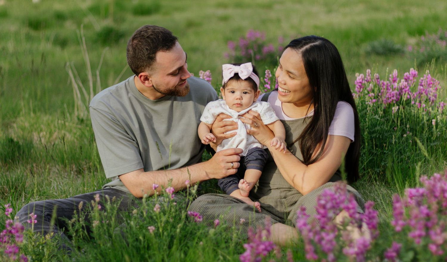 Happy young family with baby girl sitting in a lush green meadow with pink wildflowers in summer.