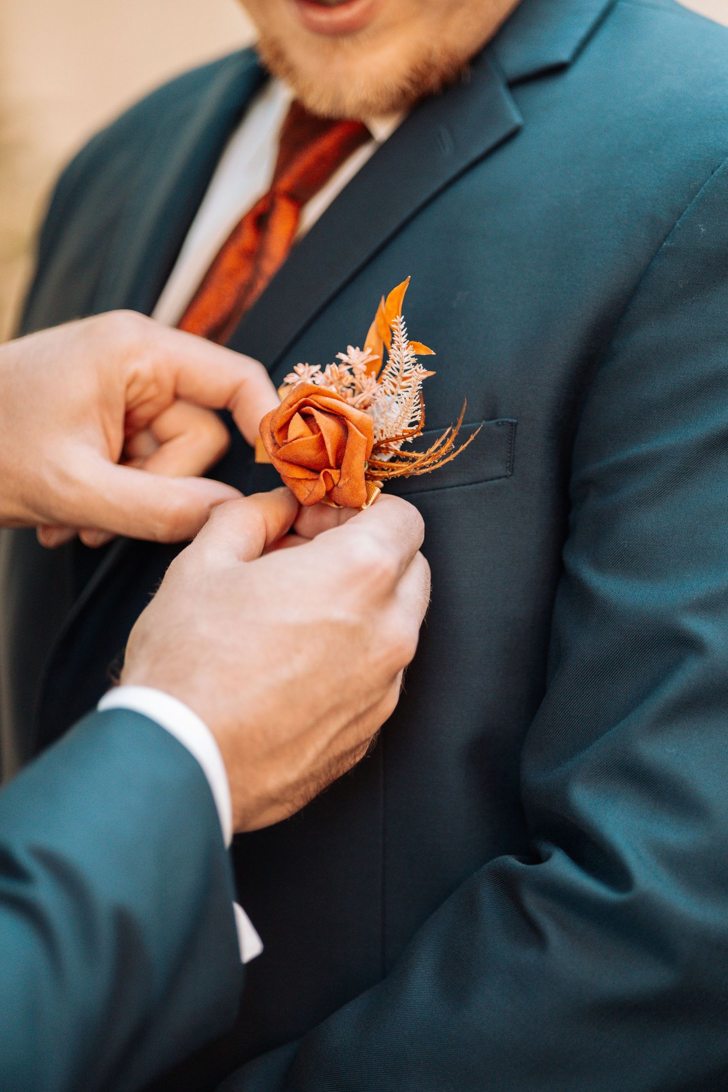 Hands pinning orange boutonniere on dark teal suit jacket.
