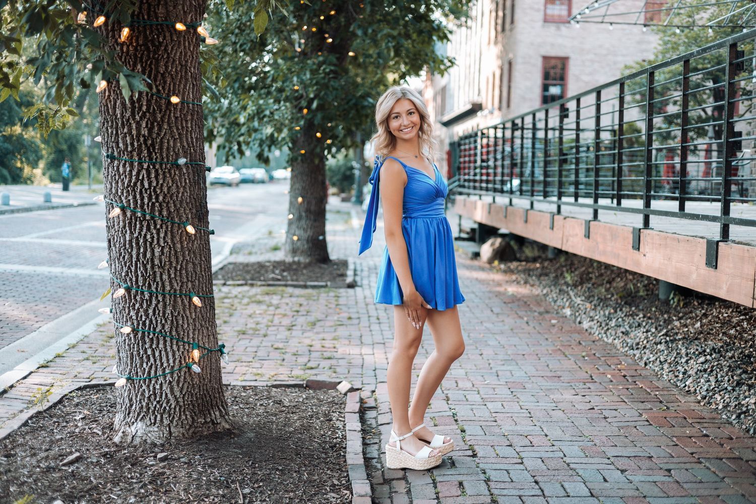 Woman in blue summer dress poses playfully on a brick sidewalk lined with trees with Kenzie Ayla Images in the Twin Cities MN area.