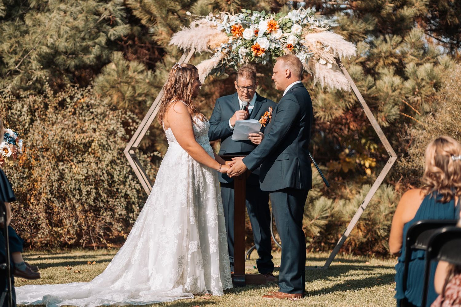 Outdoor wedding ceremony under trees with guests seated on white chairs.