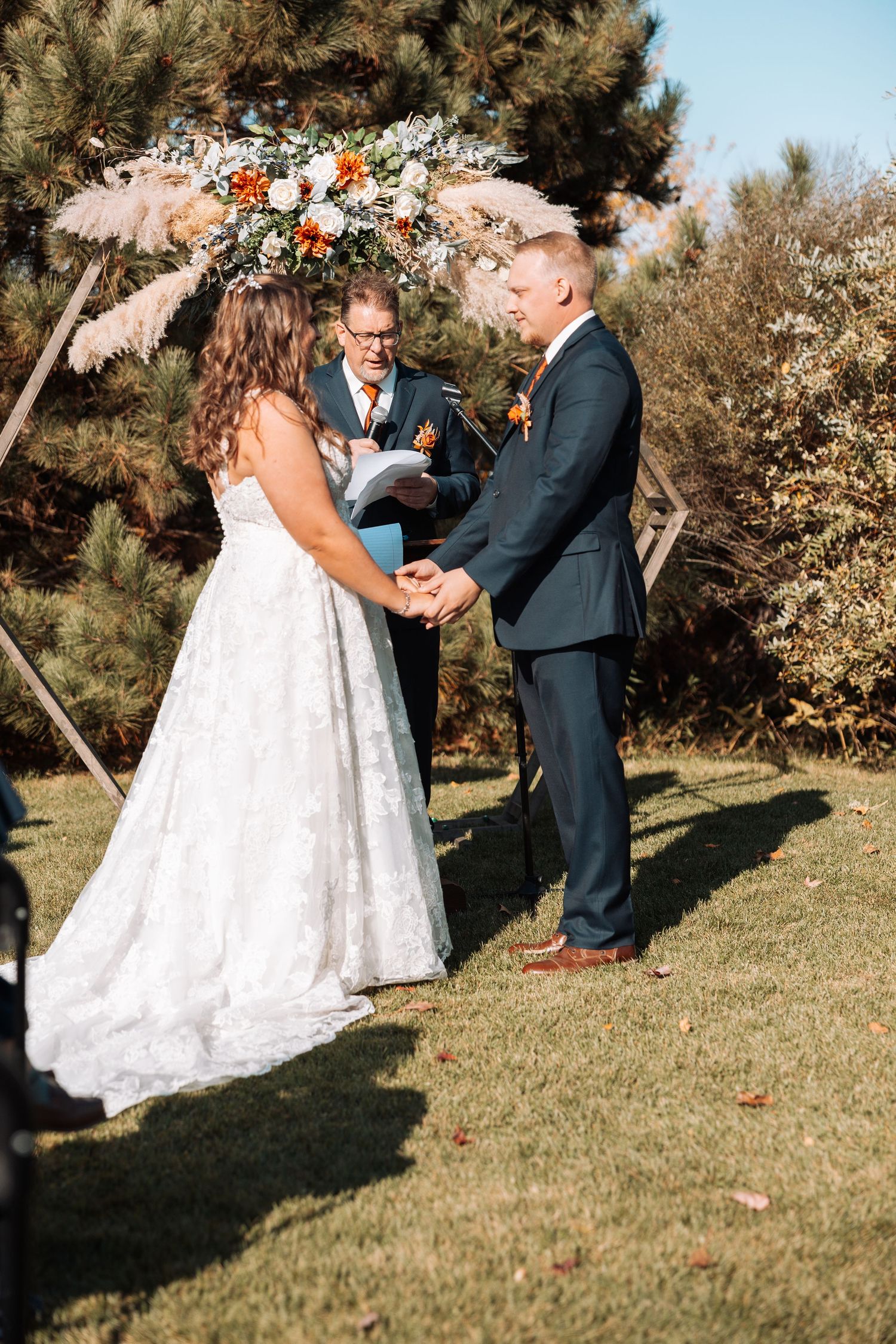 Couple exchanging vows under floral arch with pampas grass during outdoor autumn wedding.
