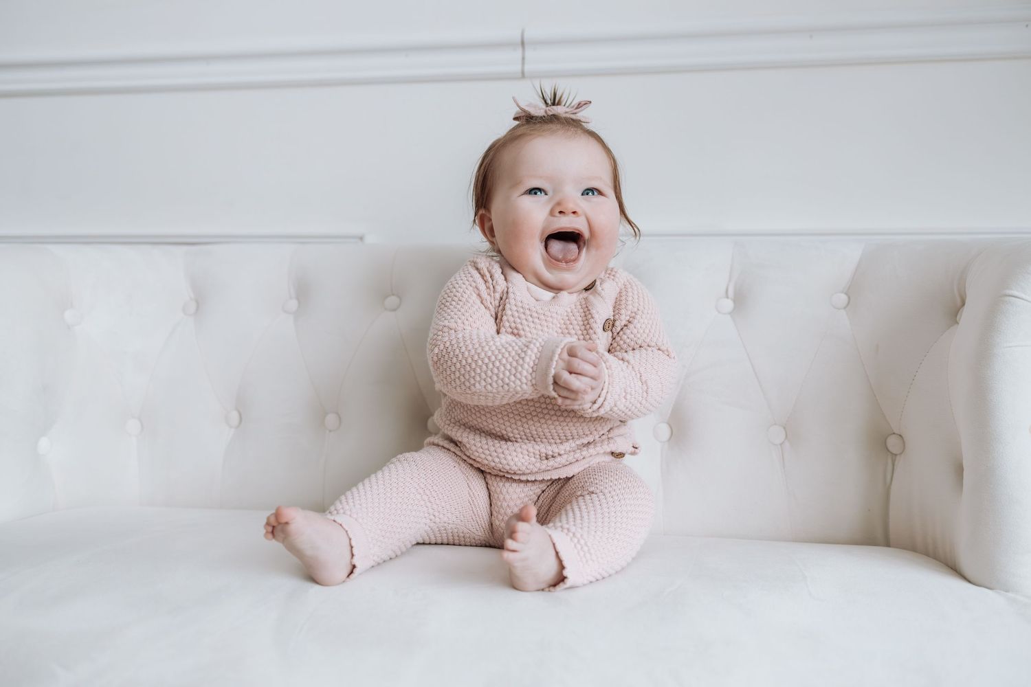 Baby in pink outfit sits happily on white tufted couch during indoor photo session.