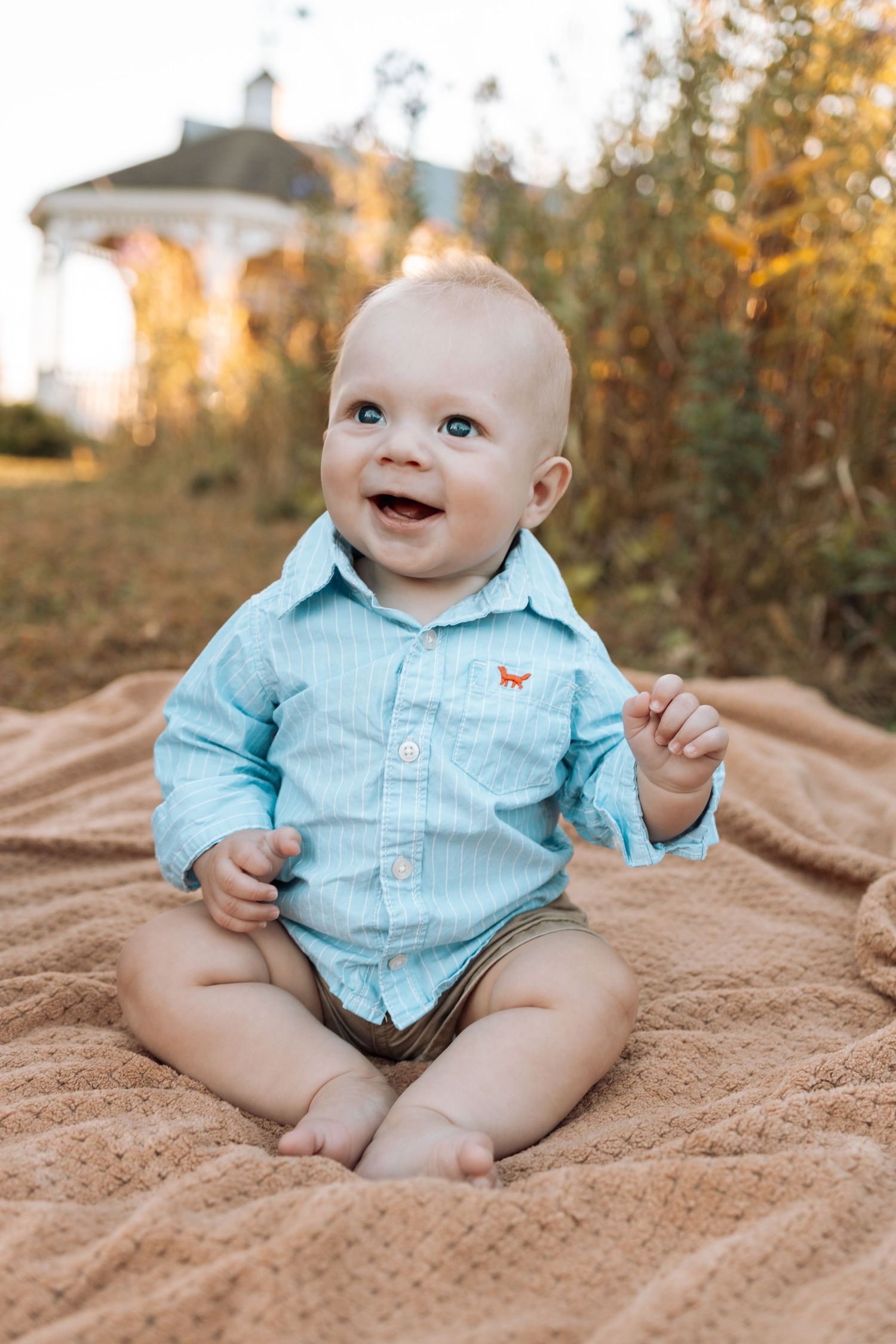 Baby wearing light blue button-up shirt sitting on textured blanket outdoors.
