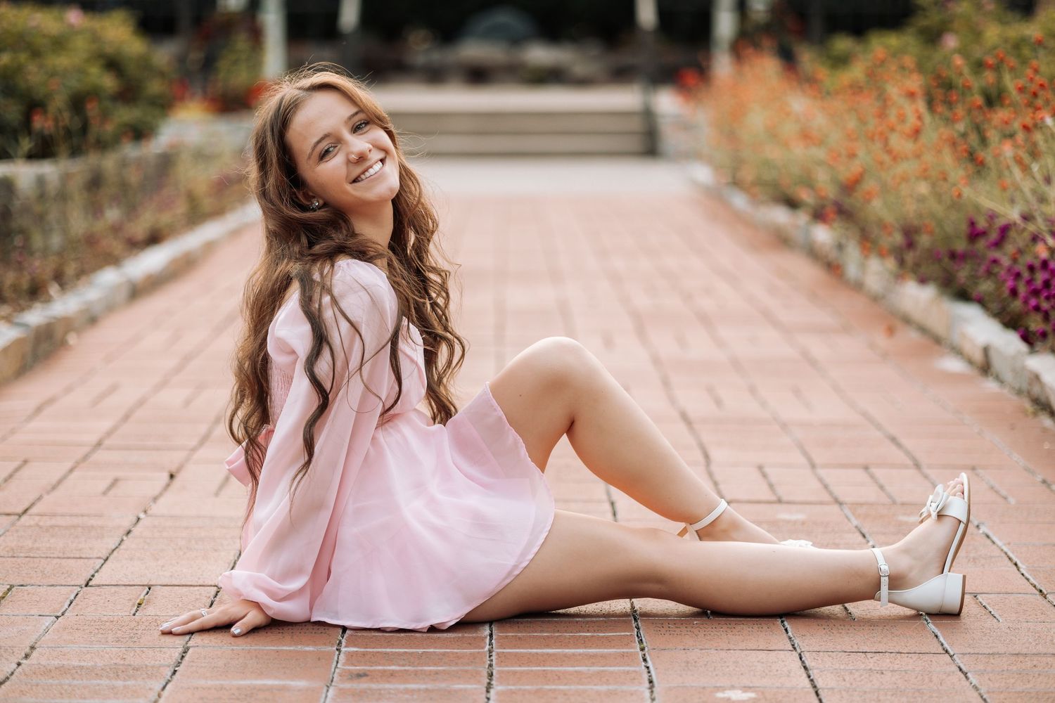 Playful senior photo session with someone in a pink dress lying on brick pavers surrounded by flowers.